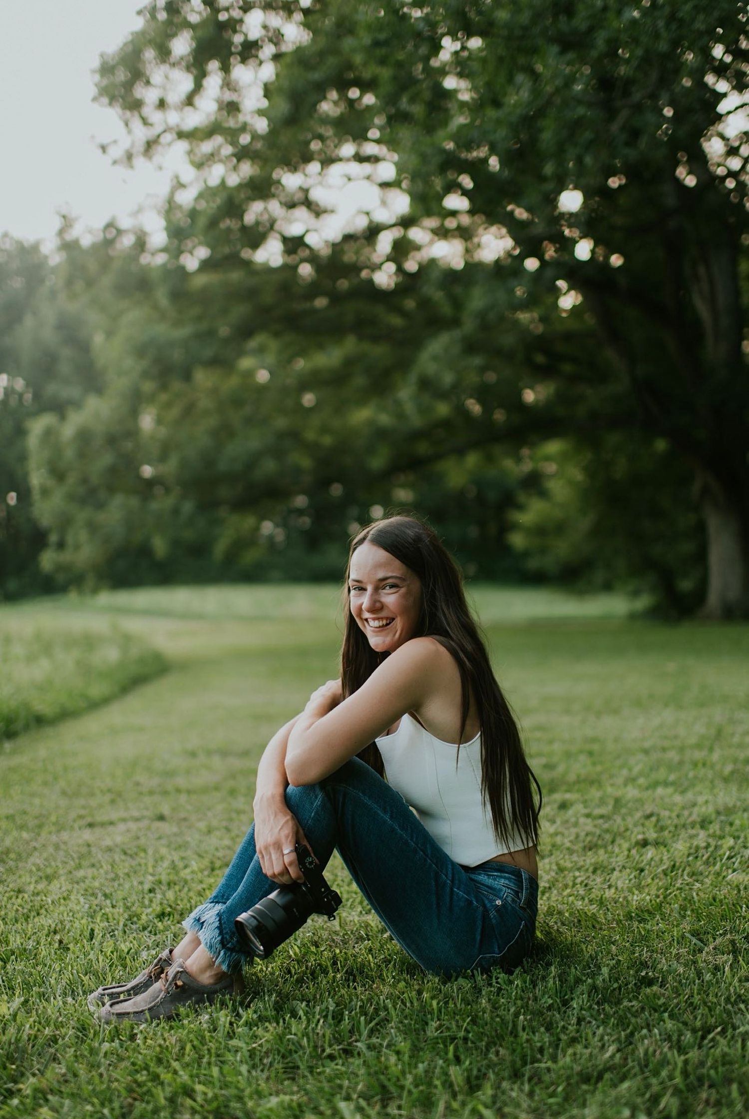 Smiling young woman with long dark hair sitting on grass in a lush green park.