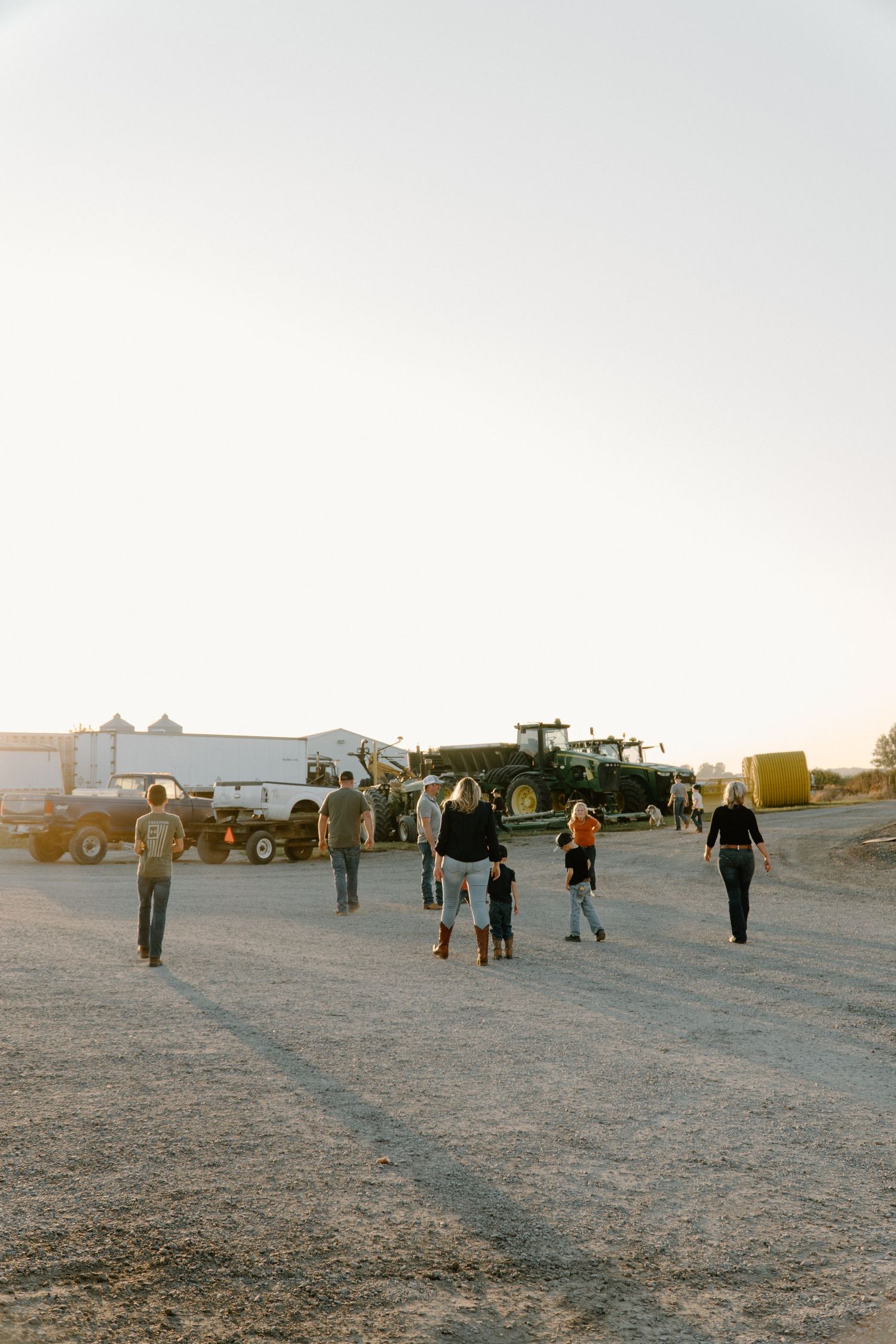 Group of people walking toward farm equipment including a green tractor at golden hour on a rural gravel lot.