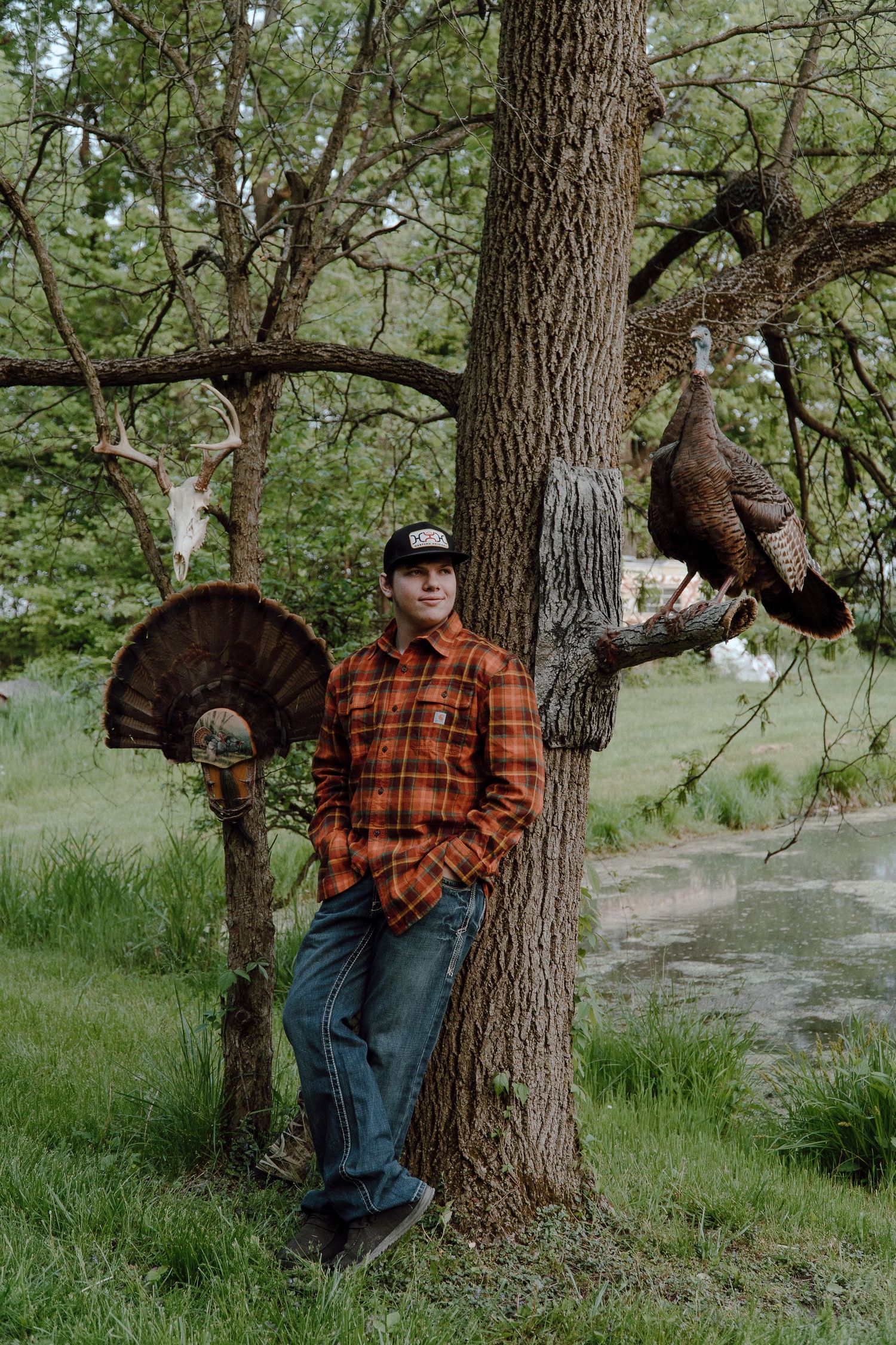 Young man in orange flannel leaning against a tree with hunted wild turkeys hanging from branches.