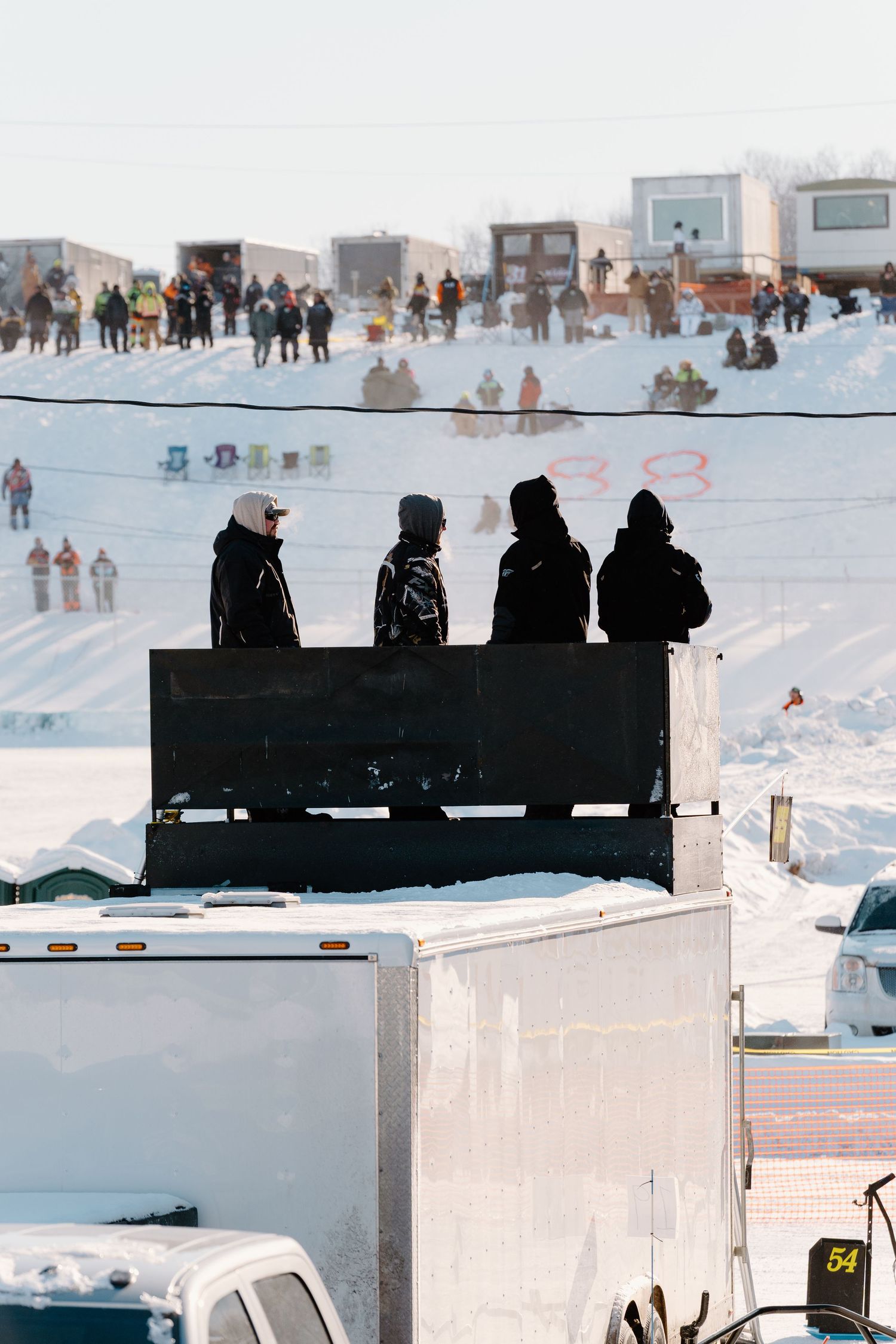 Four silhouetted spectators stand atop a trailer watching a winter snowmobile racing event on a snowy track.