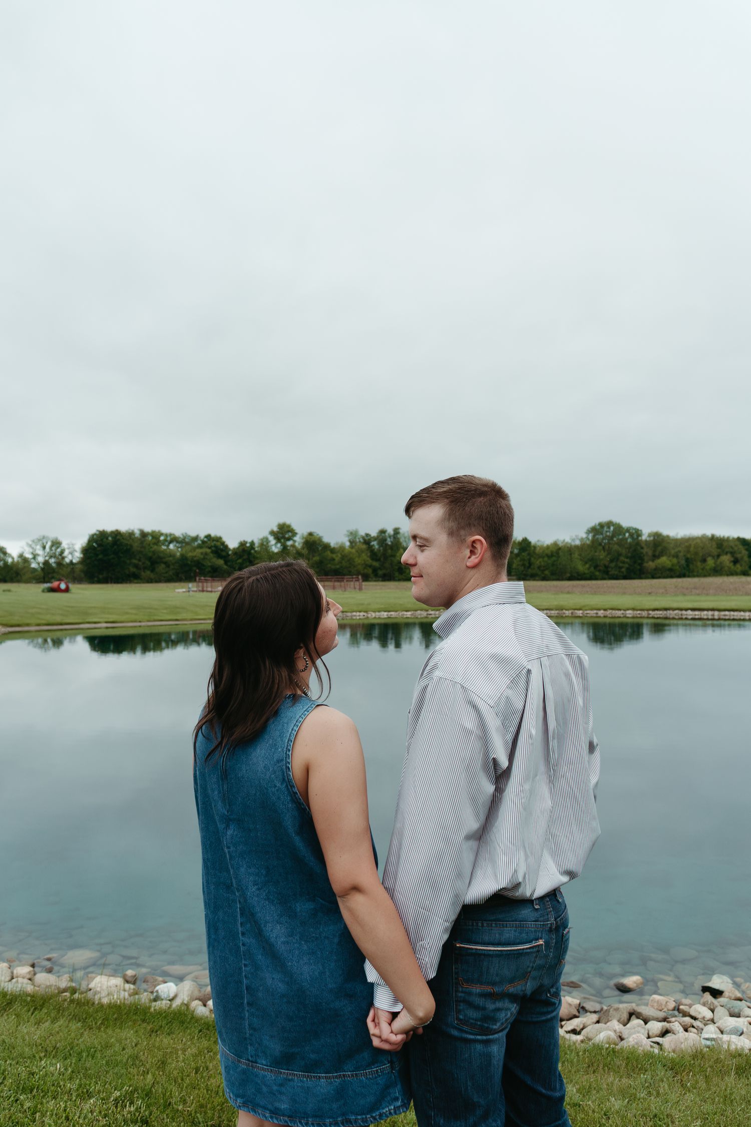 Couple holding hands while standing by a reflective pond on an overcast day in a rural setting.