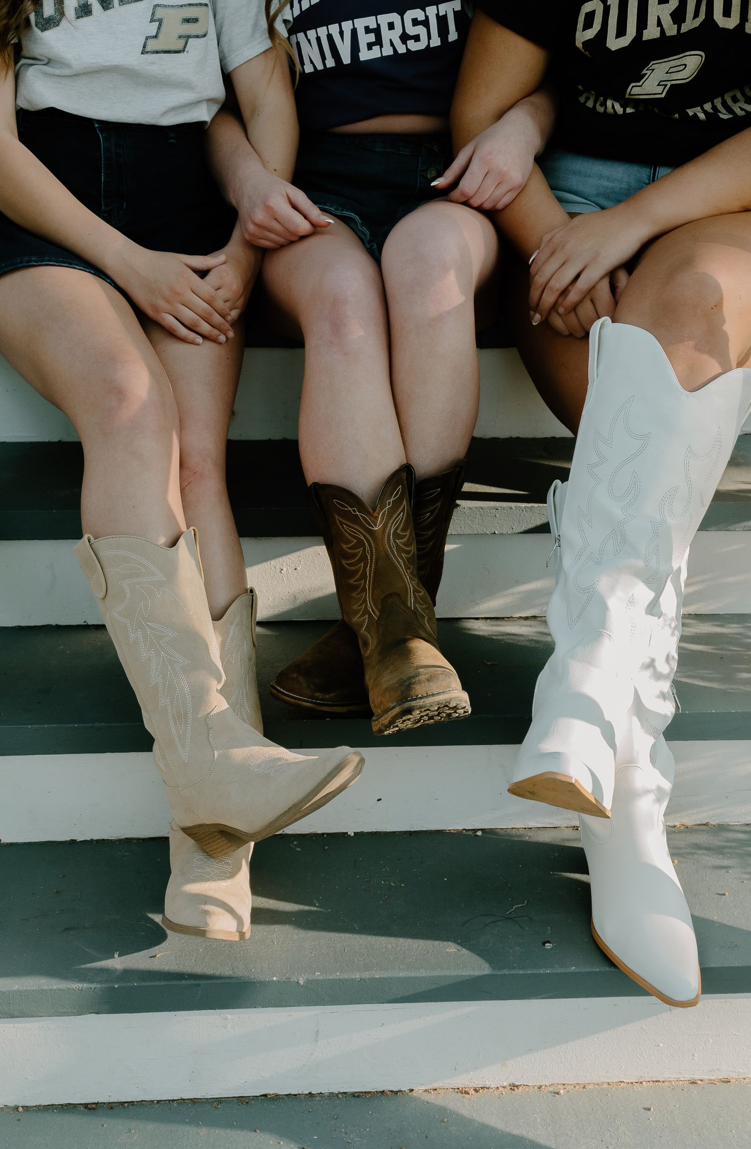 Three women in Purdue University shirts wearing cowboy boots sitting on bleachers.