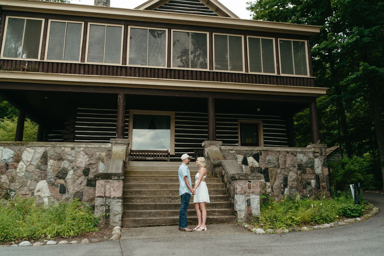 Couple holding hands on stone steps before a rustic log cabin surrounded by lush greenery.