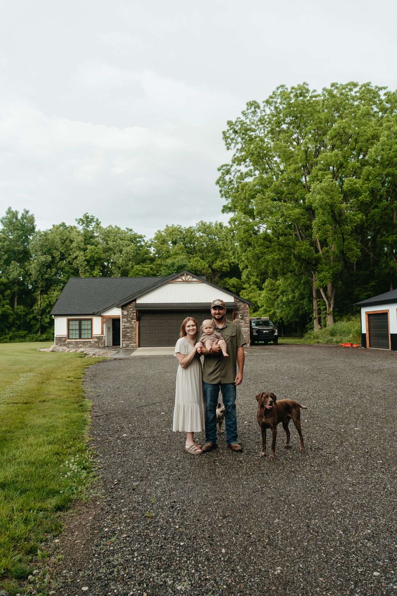 Young family with baby and brown dog stands on gravel driveway in front of ranch home.