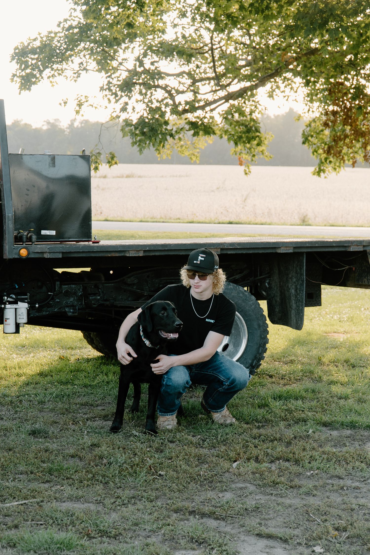 Young man with curly hair crouching beside a black lab dog near a flatbed truck in a farm field.