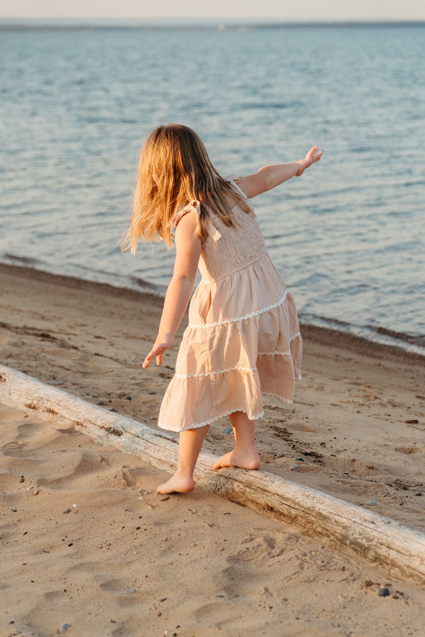 Young girl in white dress balancing on driftwood log on sandy beach near calm blue water at sunset.