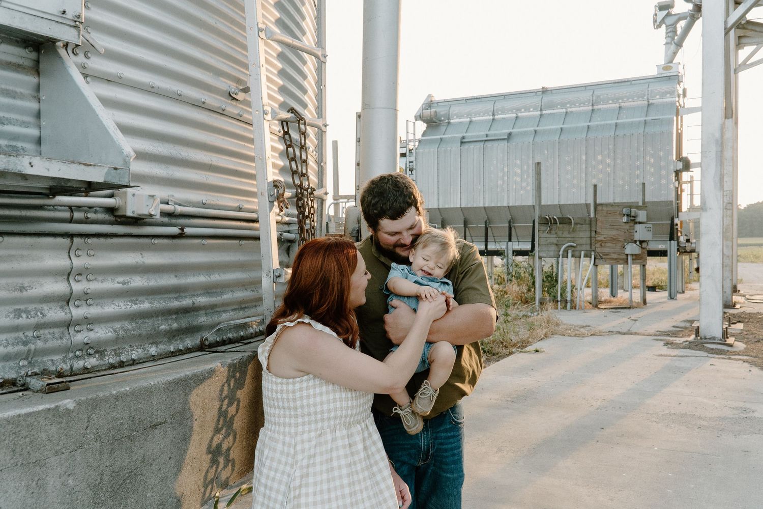 Young family with baby poses near industrial grain silos in warm golden hour light.