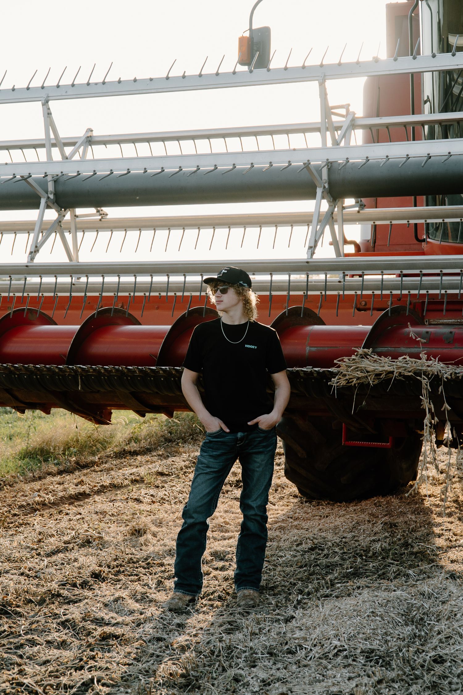 Young man in black cap and shirt standing before a red combine harvester in a harvested field.