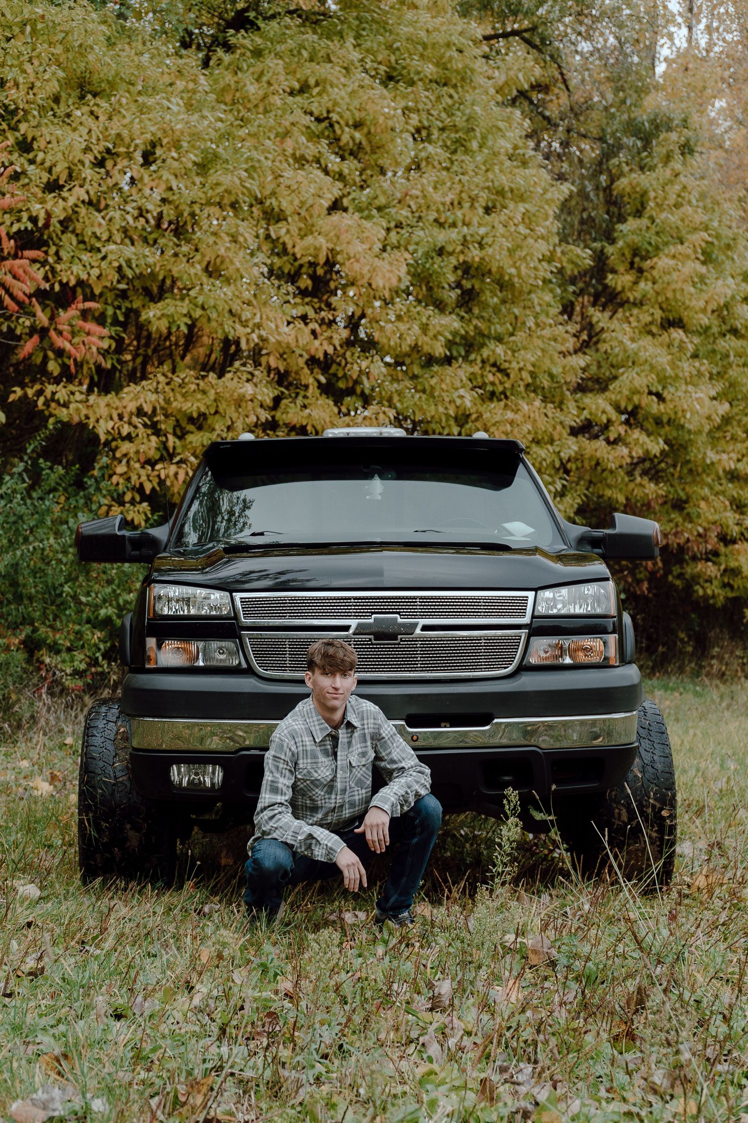 Young man in plaid shirt crouching in front of a lifted black Chevrolet truck in autumn woods.