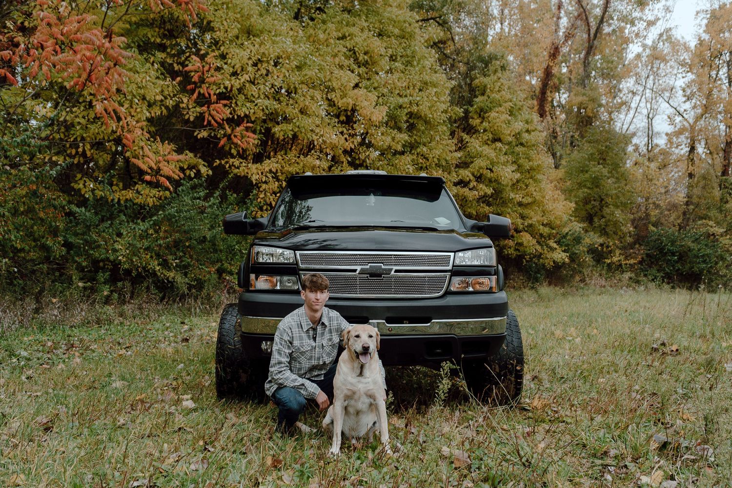 Young man kneeling beside a yellow Labrador dog in front of a lifted black Chevrolet truck outdoors.