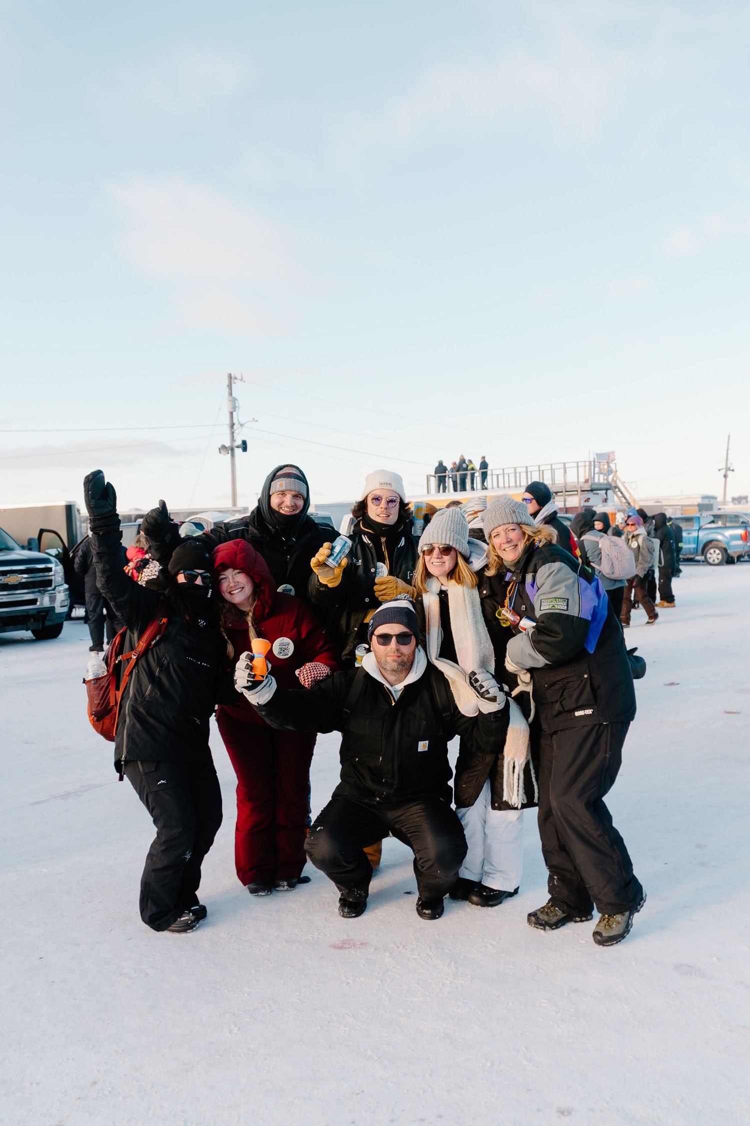 Group of seven friends posing together in winter gear holding drinks at an outdoor snowmobile racing event.