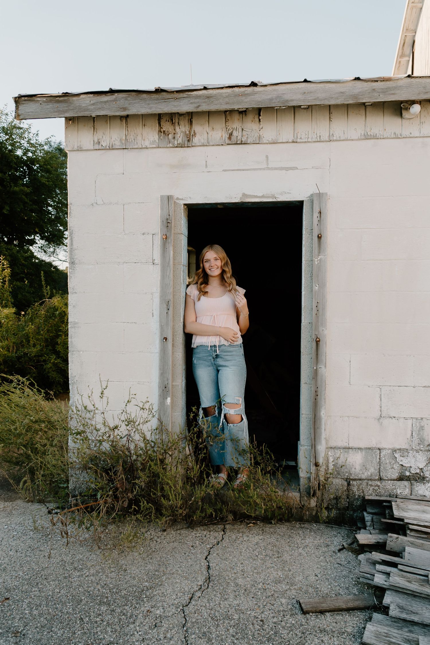 Young woman in pink top and ripped jeans smiling in doorway of rustic white concrete building.