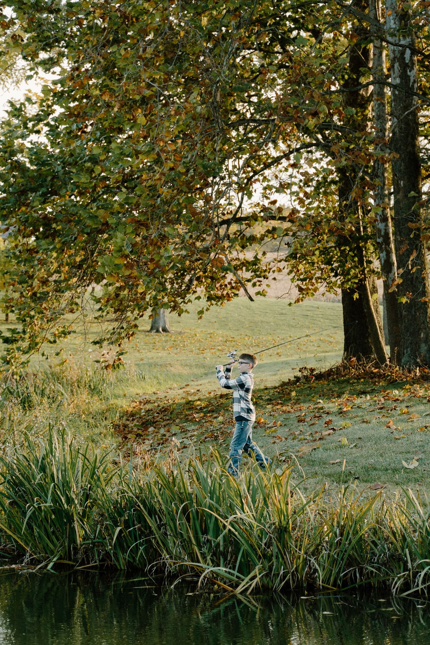 Young boy fishing with a rod along a pond bank under large autumn trees.