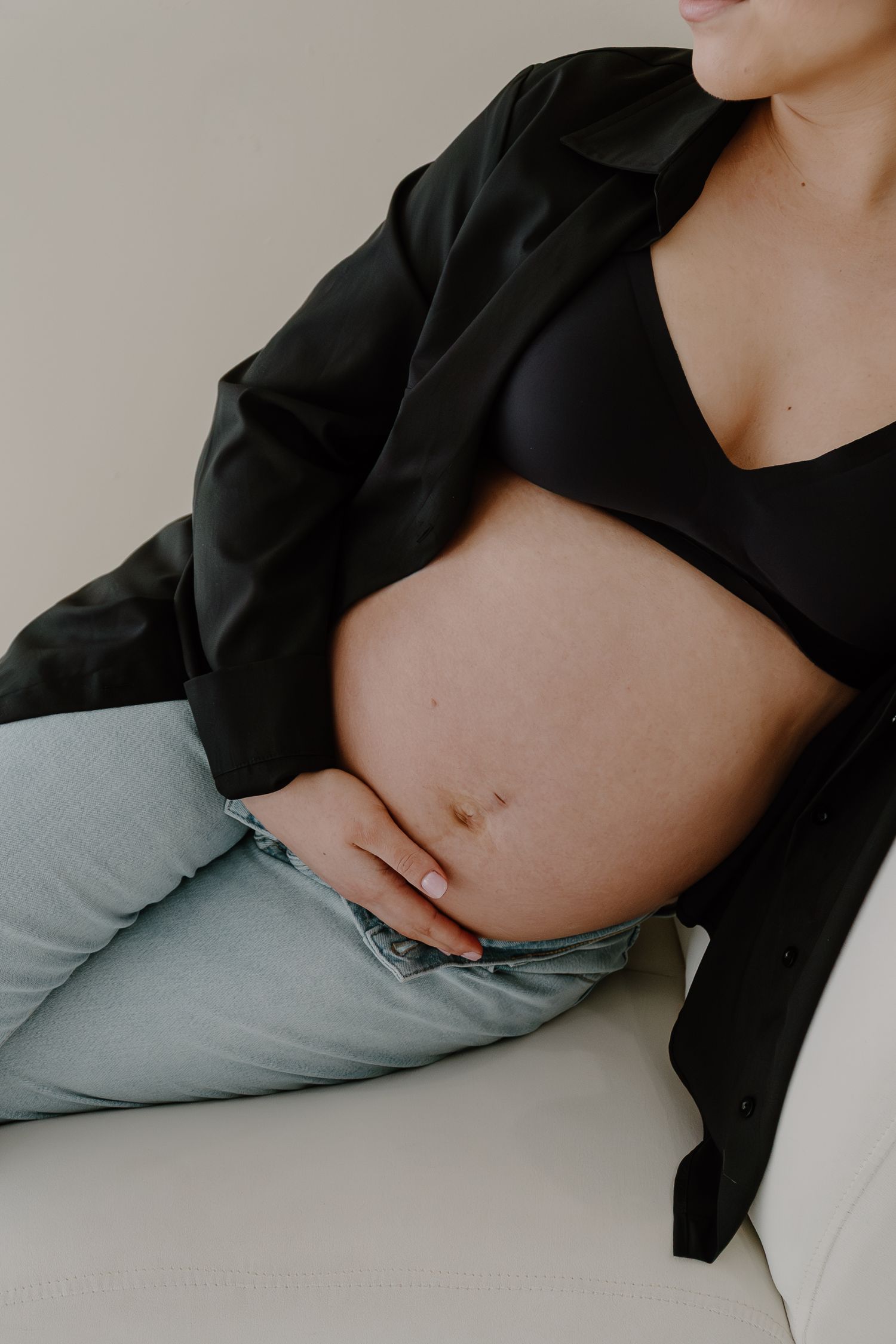Close-up of pregnant woman in black jacket and jeans cradling bare baby bump.