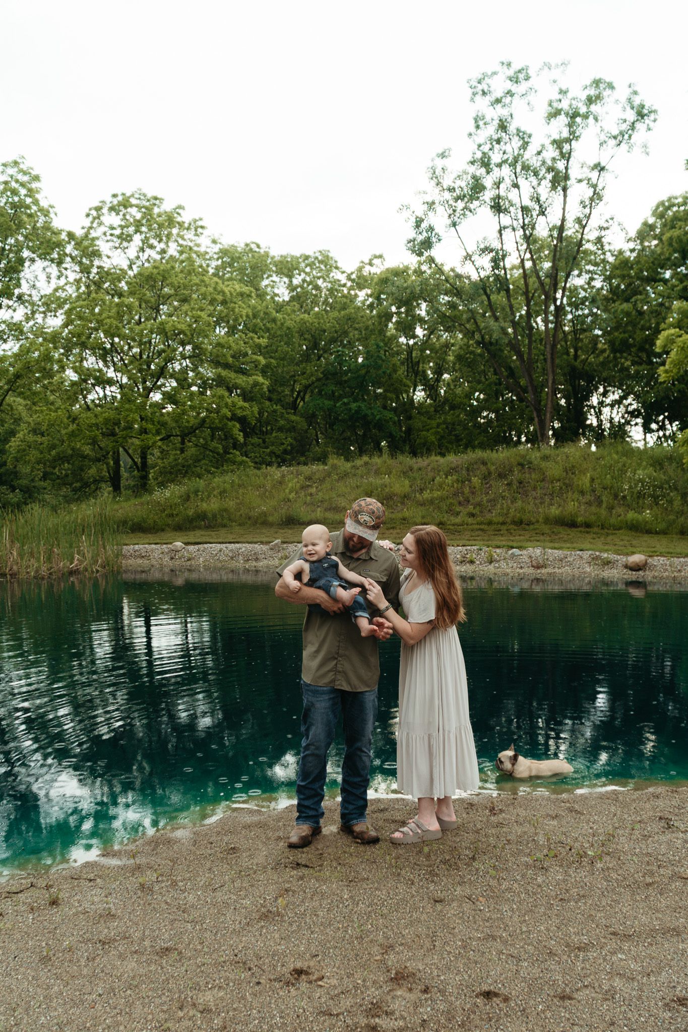 Young family with baby poses together beside a serene blue-green pond surrounded by lush green trees.