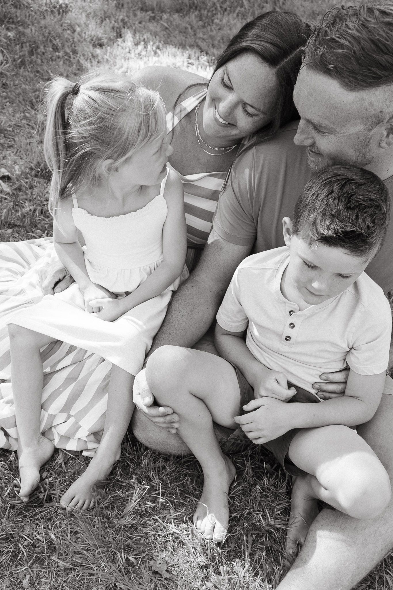 Black and white overhead photo of happy family of four sitting closely together on grass outdoors.