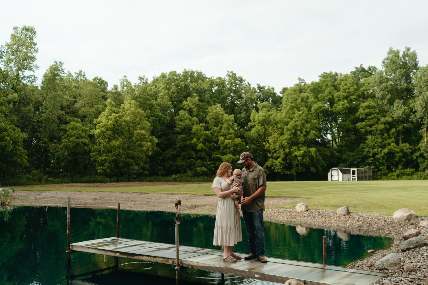 Couple holding a baby stands on a wooden dock overlooking a calm blue-green pond with green trees behind.