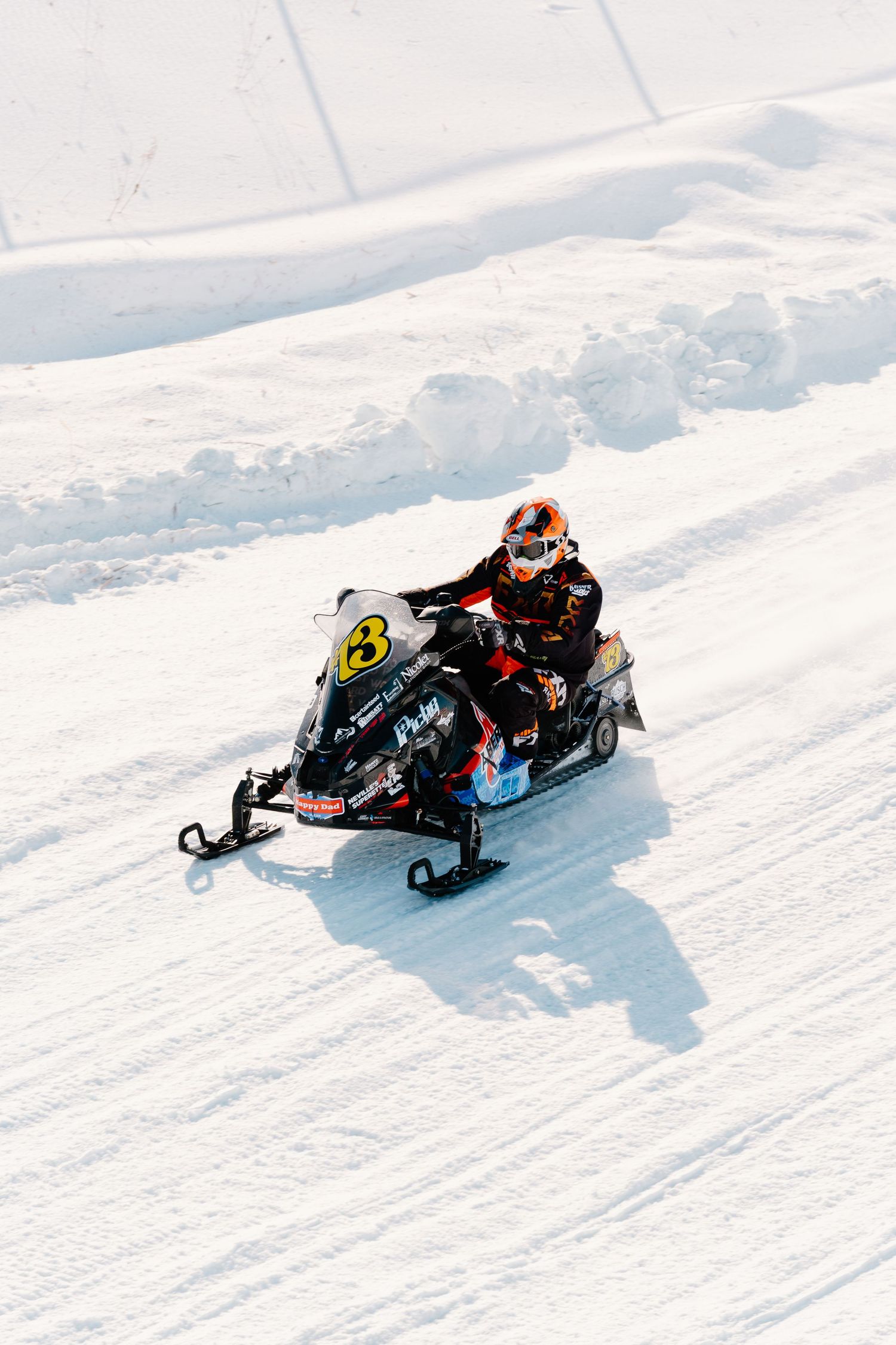 Snowmobile racer wearing orange helmet riding black sled number 3 on a bright snowy race track.