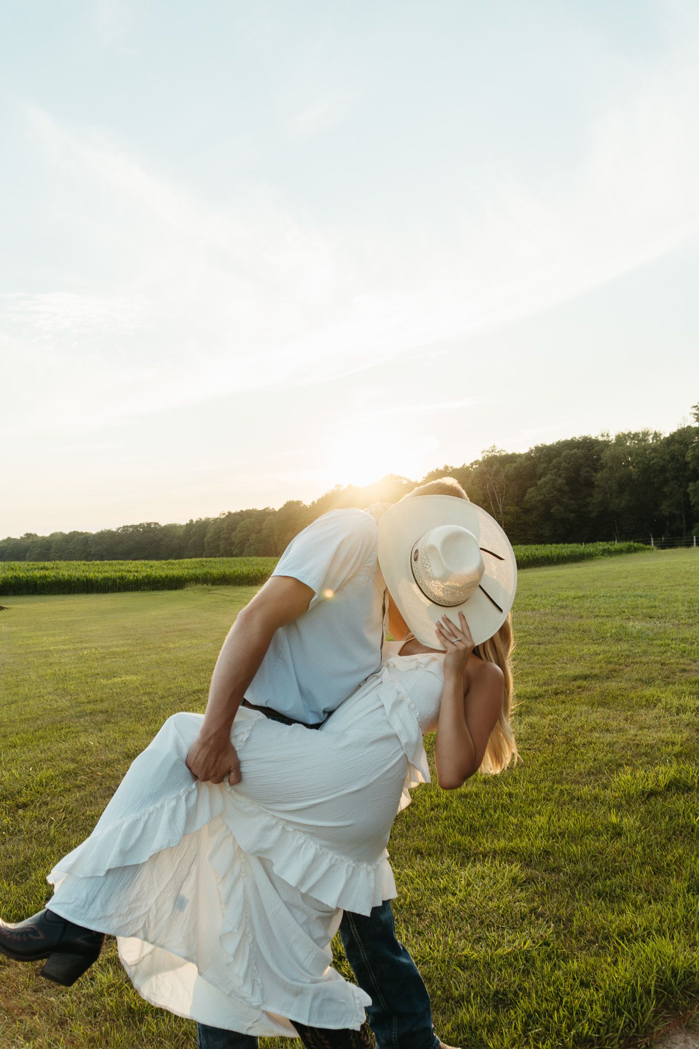 Man dipping woman in white dress for a kiss at golden sunset in an open green field.