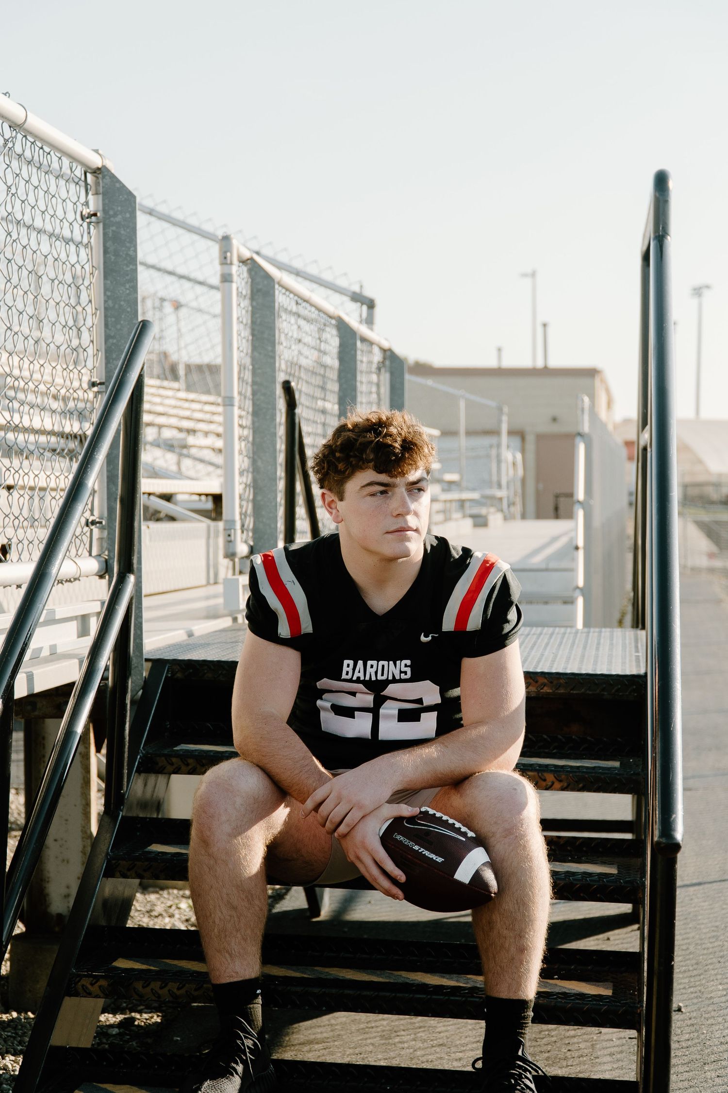 Teen athlete in Barons number 22 football jersey sitting on stadium stairs holding a football outdoors.