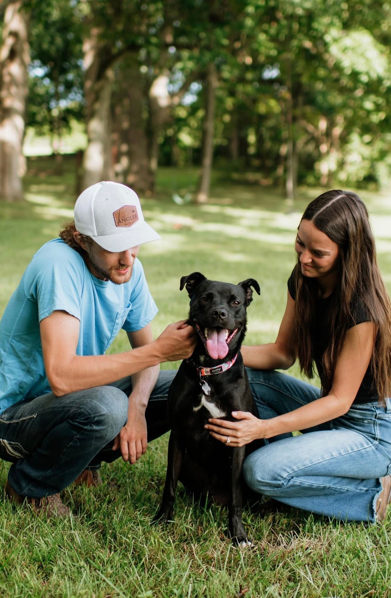 Couple crouching in grass petting their happy black dog in a sunny park.