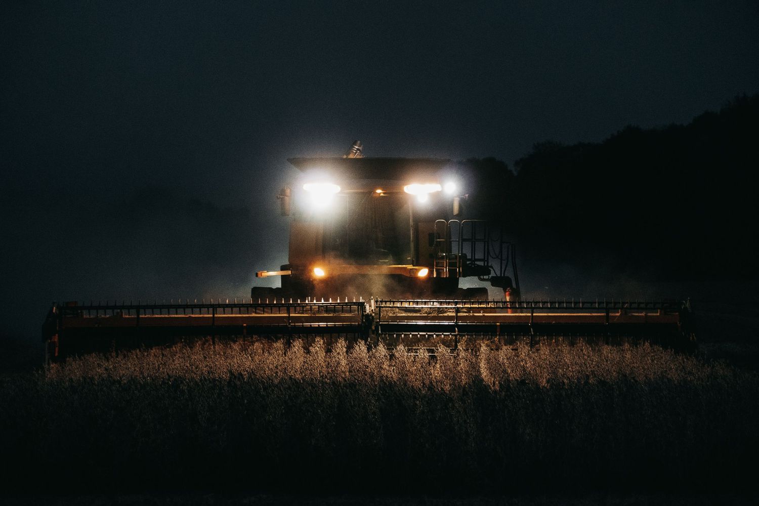 Agricultural combine harvester working in a field at night with bright headlights illuminating crops.