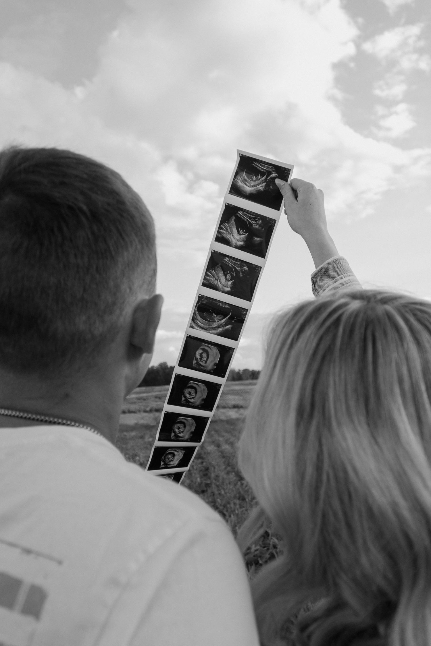 Black and white image of couple viewing ultrasound strip photos against cloudy sky.