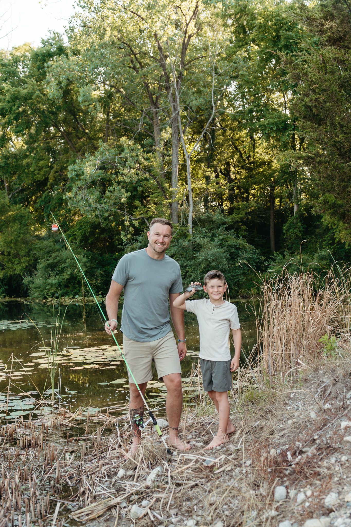 A father and son stand together smiling by a peaceful pond holding fishing rods in a wooded area.