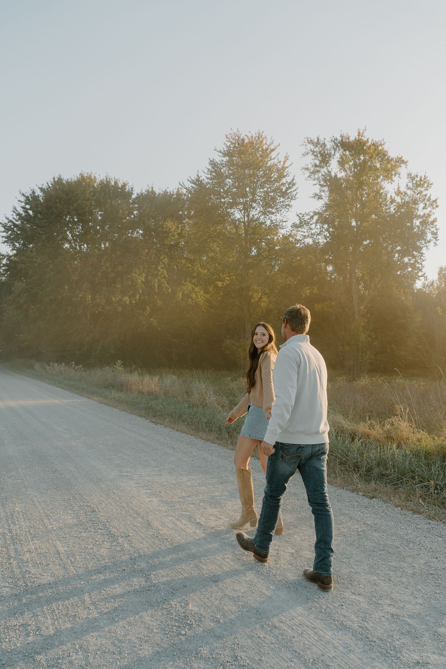 Couple holding hands walking on a sunny rural dirt road surrounded by golden trees at sunset.