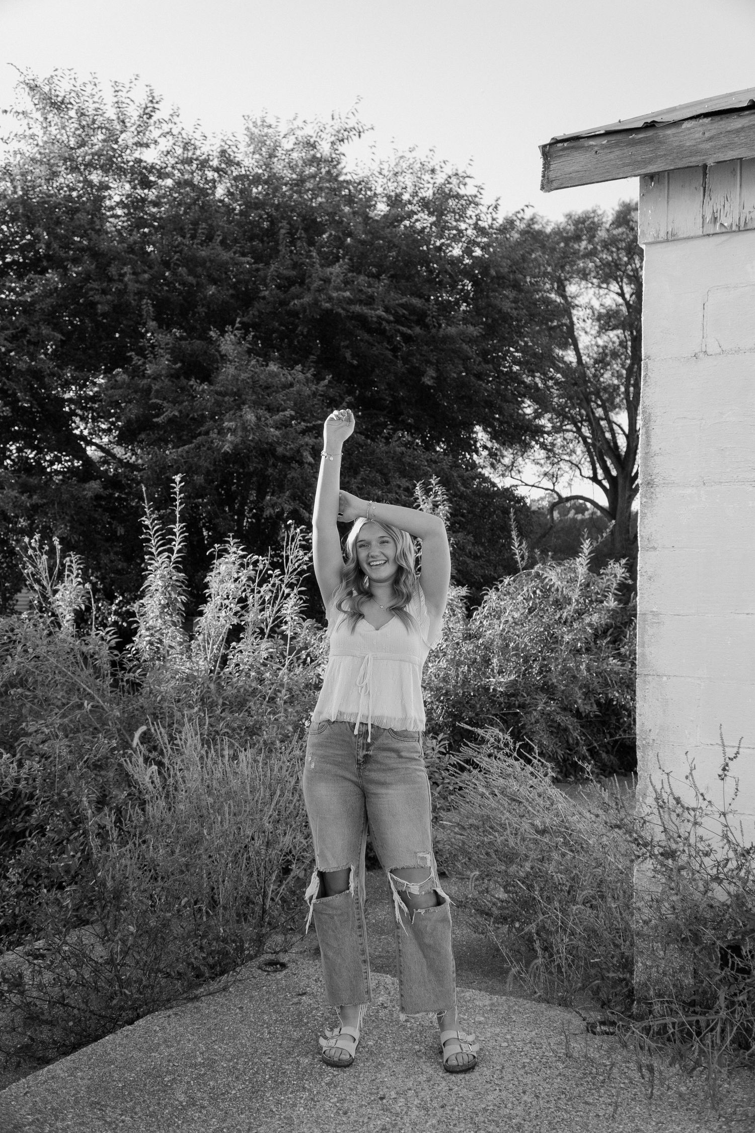 Black and white photo of a smiling young woman raising her arm near an overgrown abandoned building.