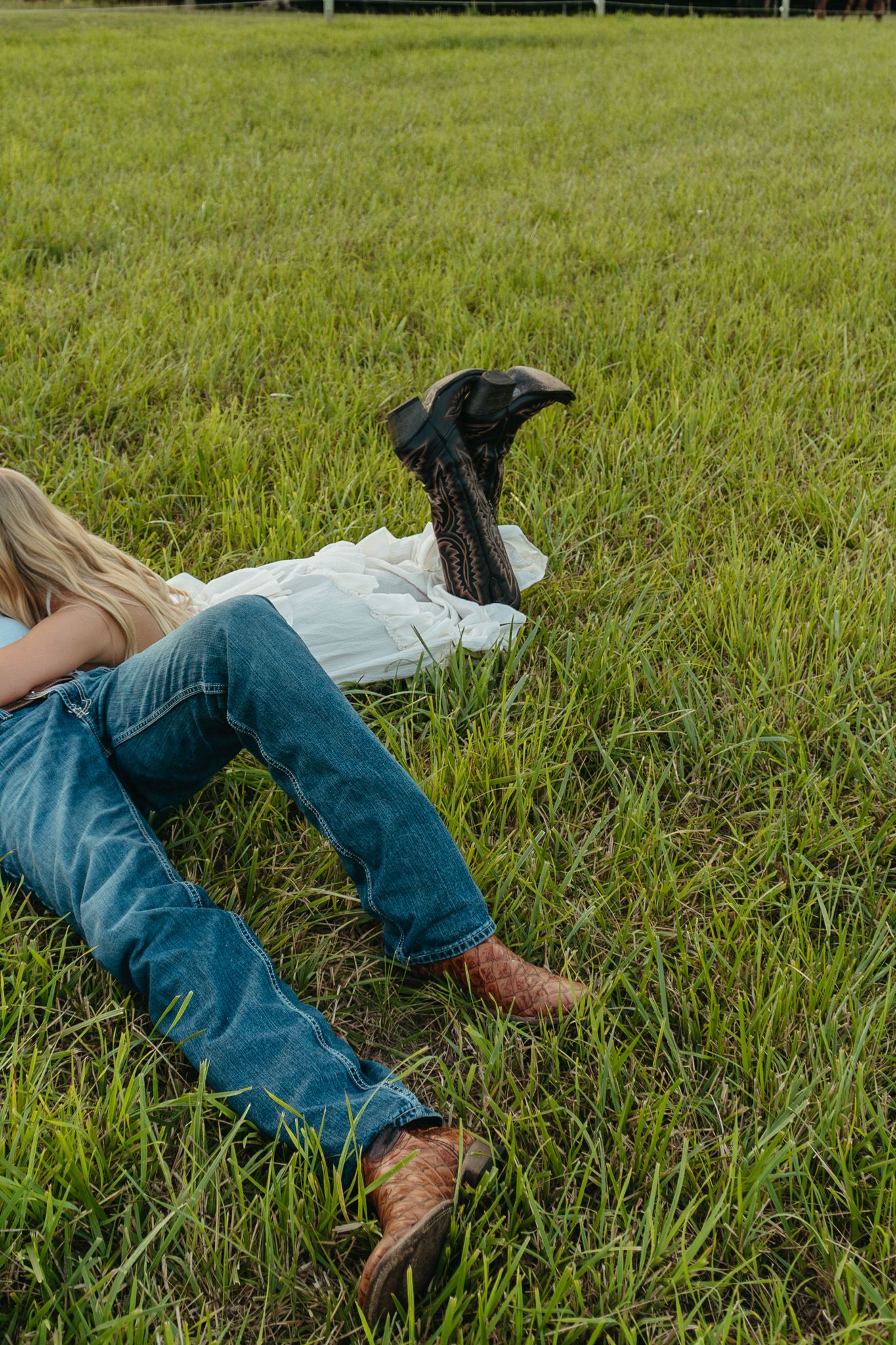 Two people lying in green grass wearing jeans and cowboy boots on a sunny day.