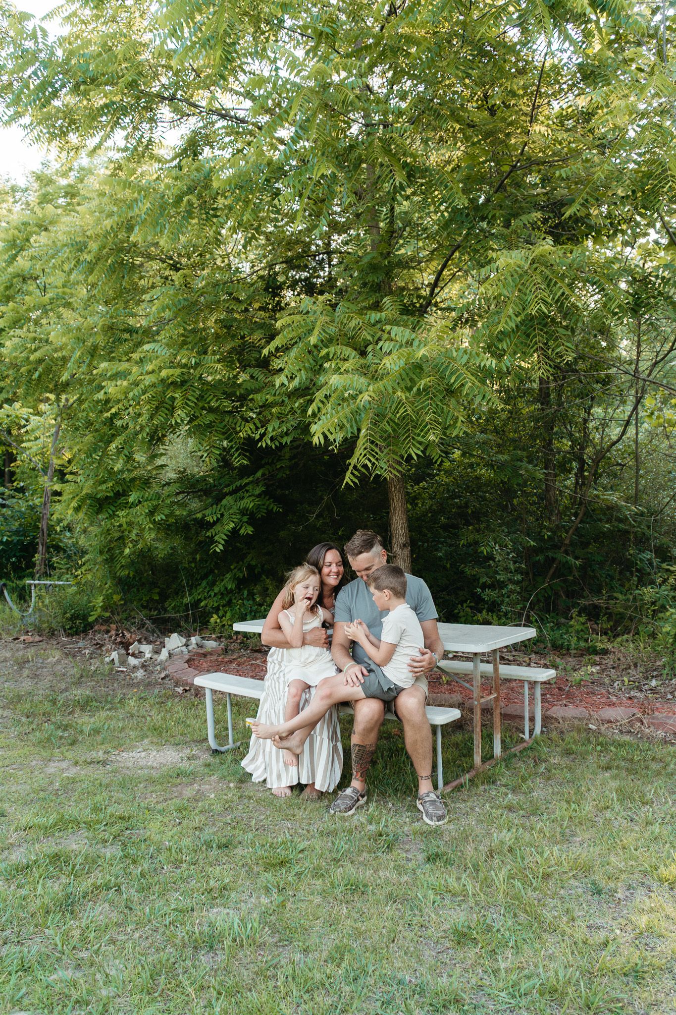 A loving family of four sits closely together at a picnic table under a large green tree.
