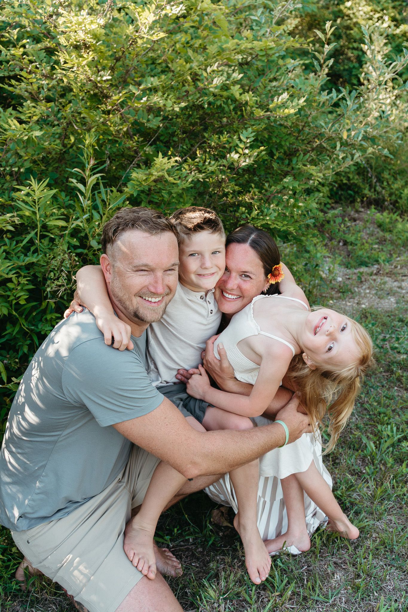 A happy family of four hugs and smiles together outdoors surrounded by lush green foliage.