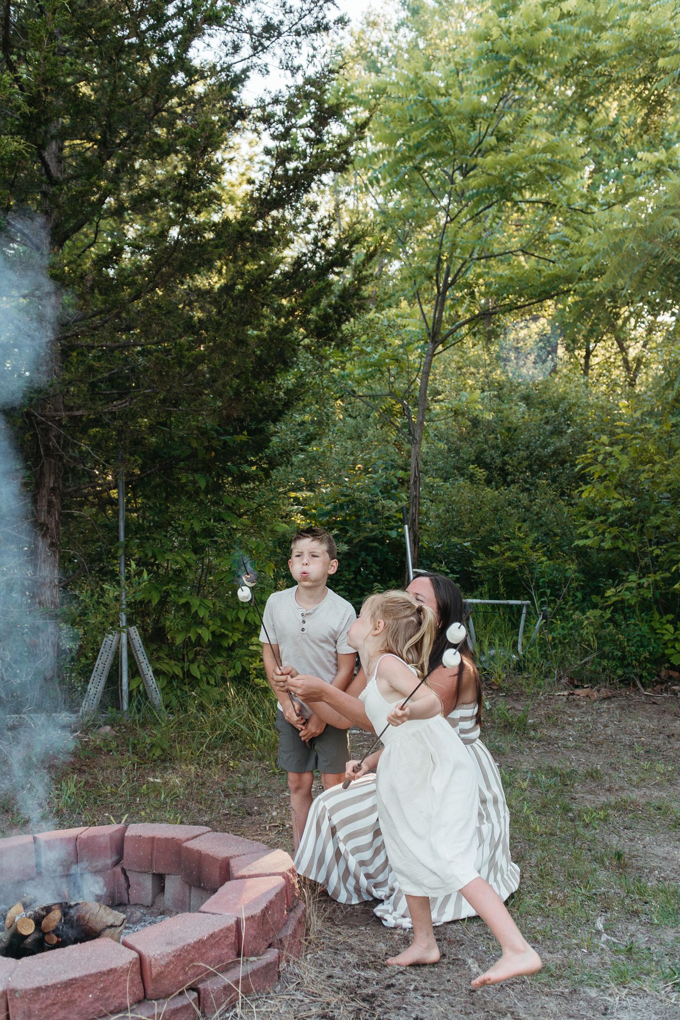 Children roast marshmallows over a backyard brick fire pit surrounded by green trees at dusk.