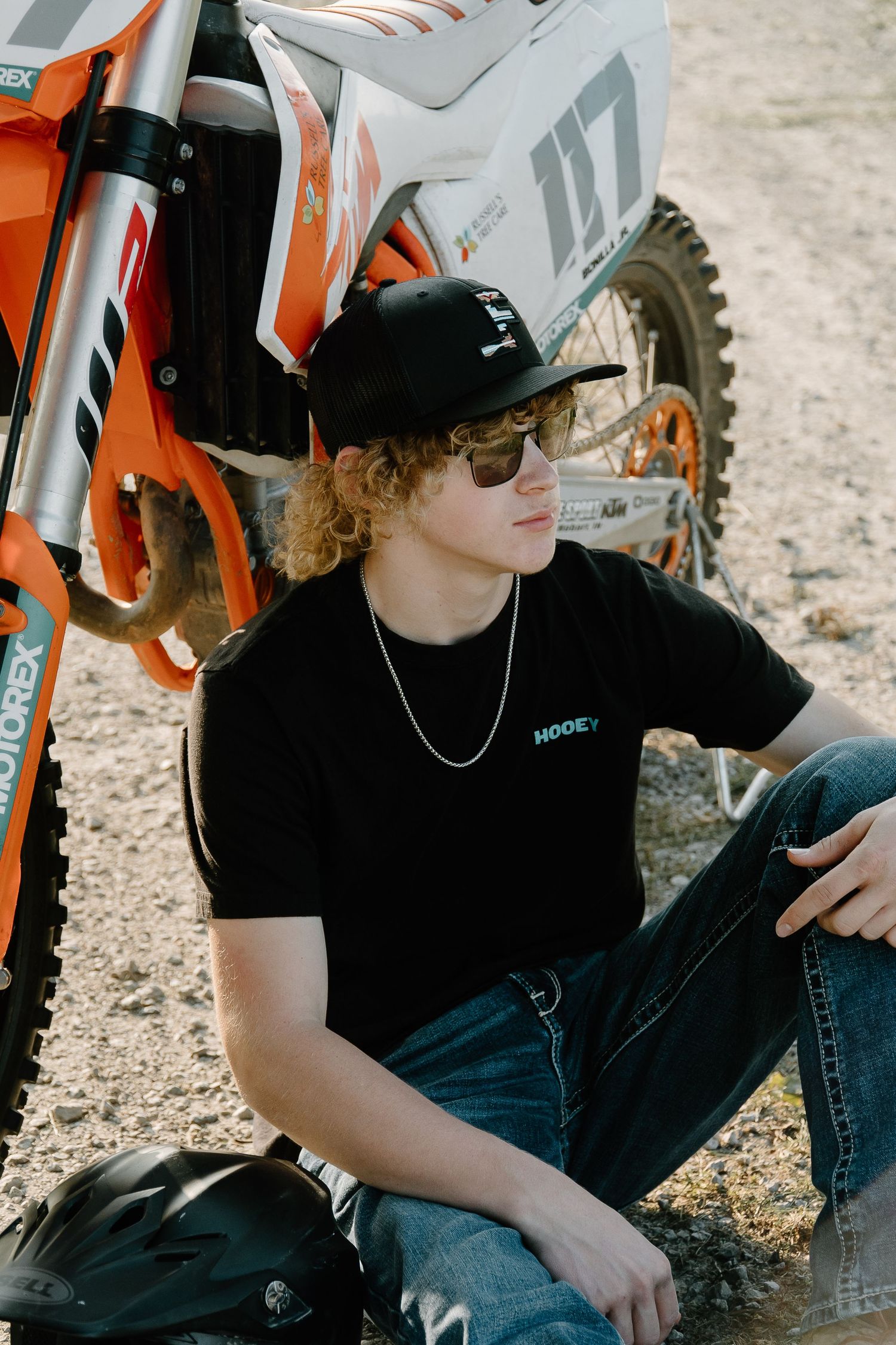Young man with curly hair sitting beside an orange KTM dirt bike wearing a black Hooey shirt and cap.