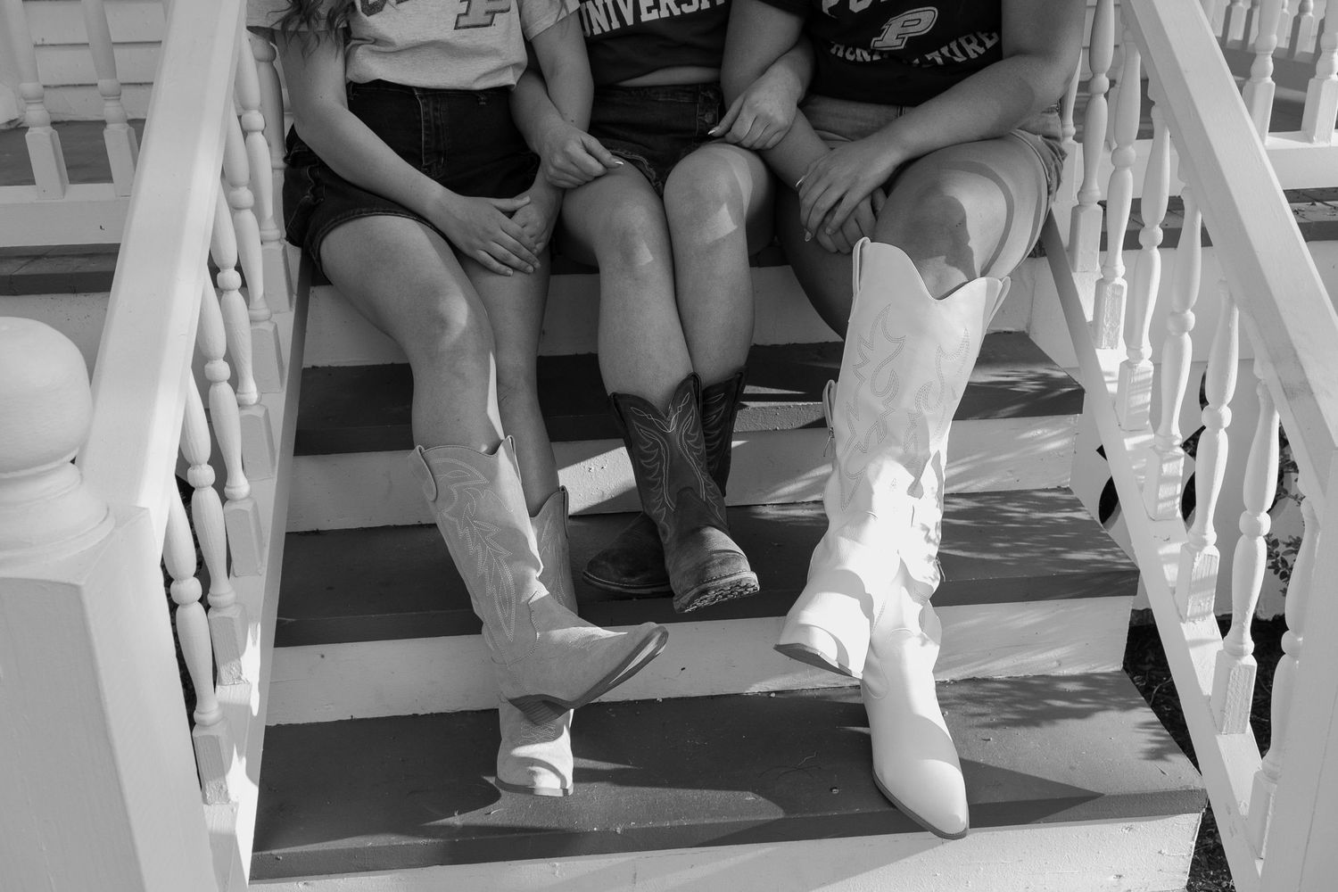 Three people sitting on porch steps wearing cowboy boots in a black and white photo.