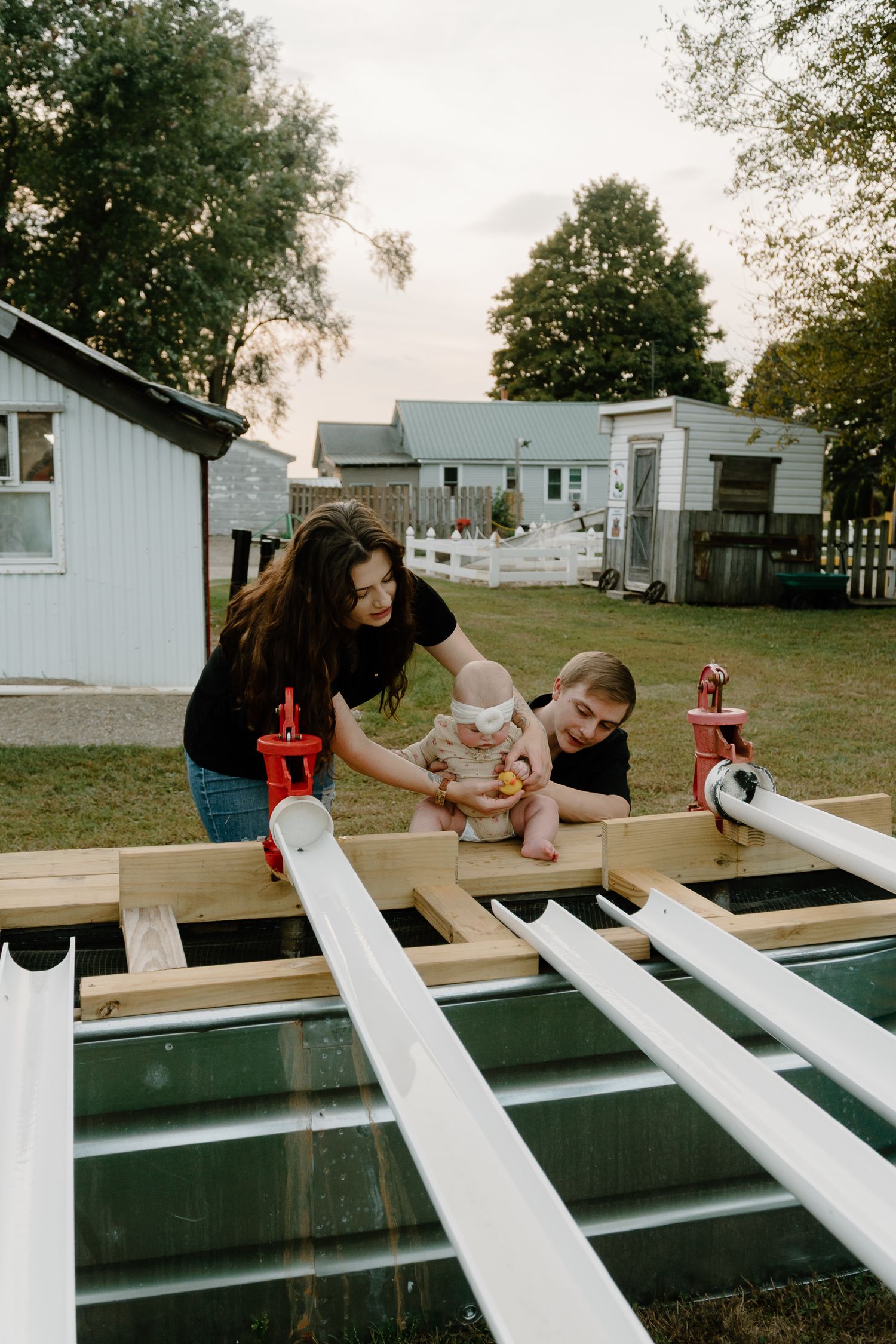 Young couple with baby working on a woodworking project outdoors in backyard at dusk near white outbuildings.