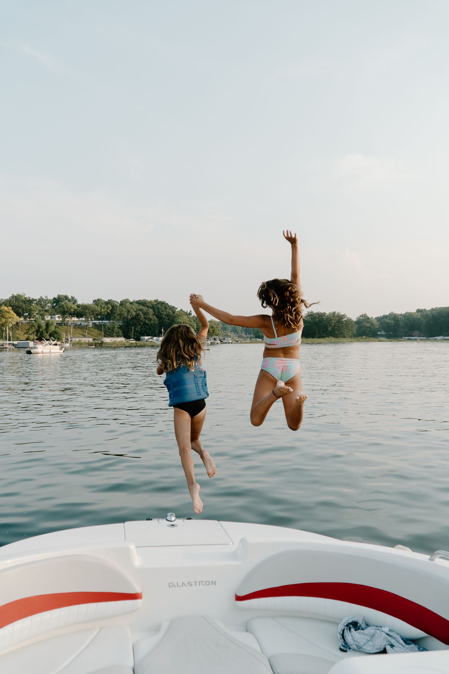 Two girls jumping off white boat into calm lake at sunset.