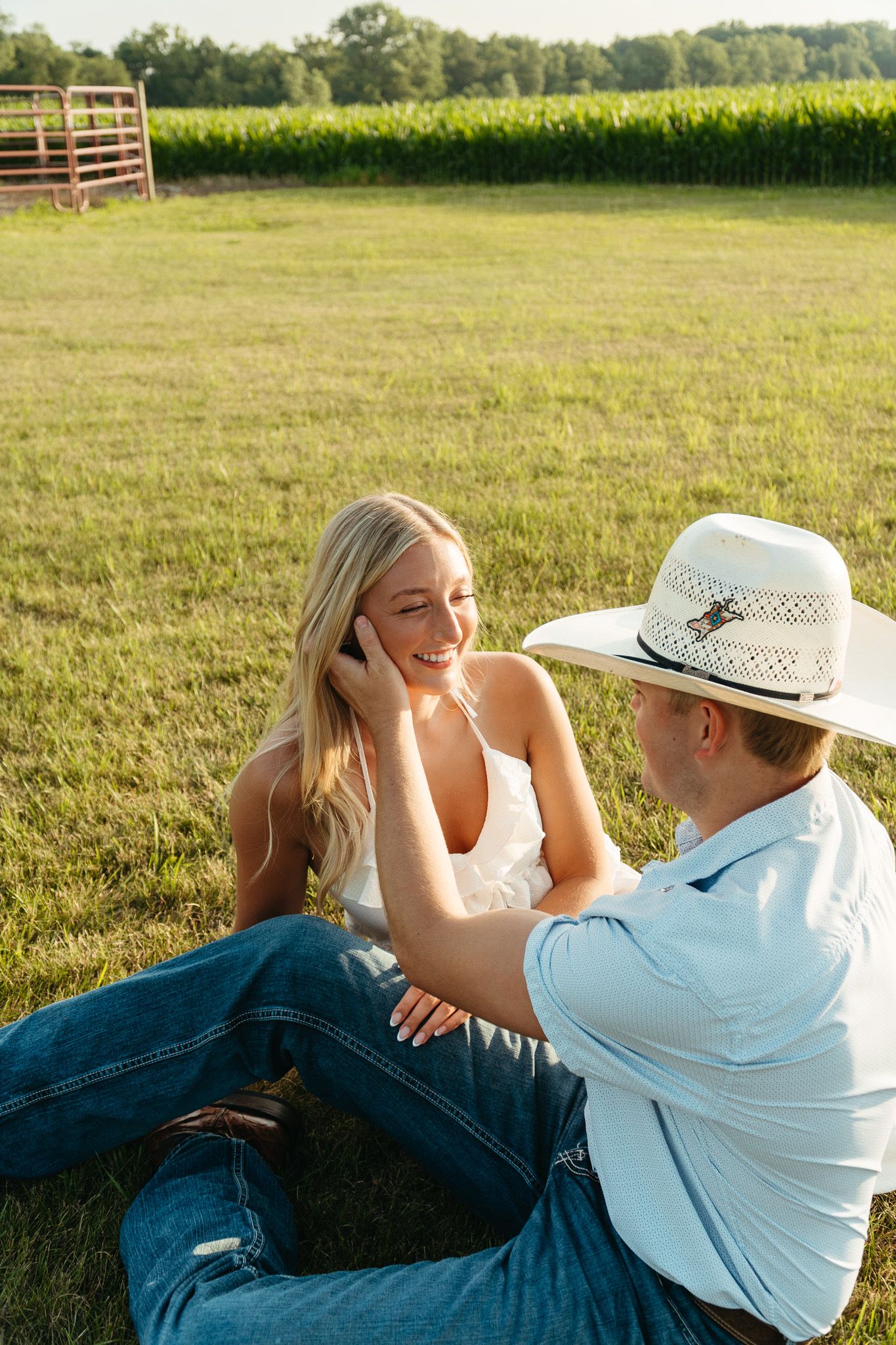 Couple sitting in sunny green field, woman laughing as man in cowboy hat reaches toward her hair.