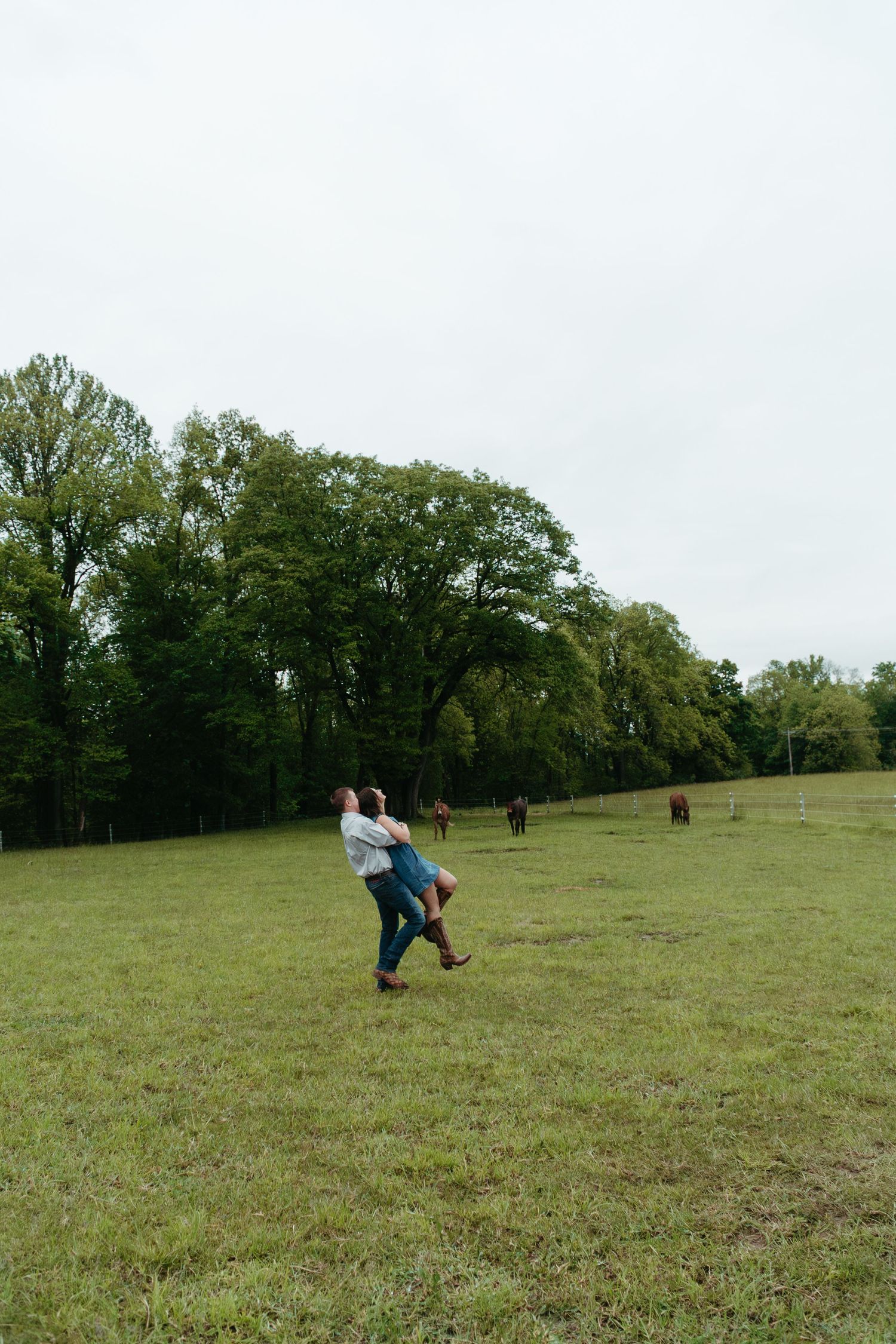 Couple spinning joyfully in a wide green pasture with horses grazing in the background.