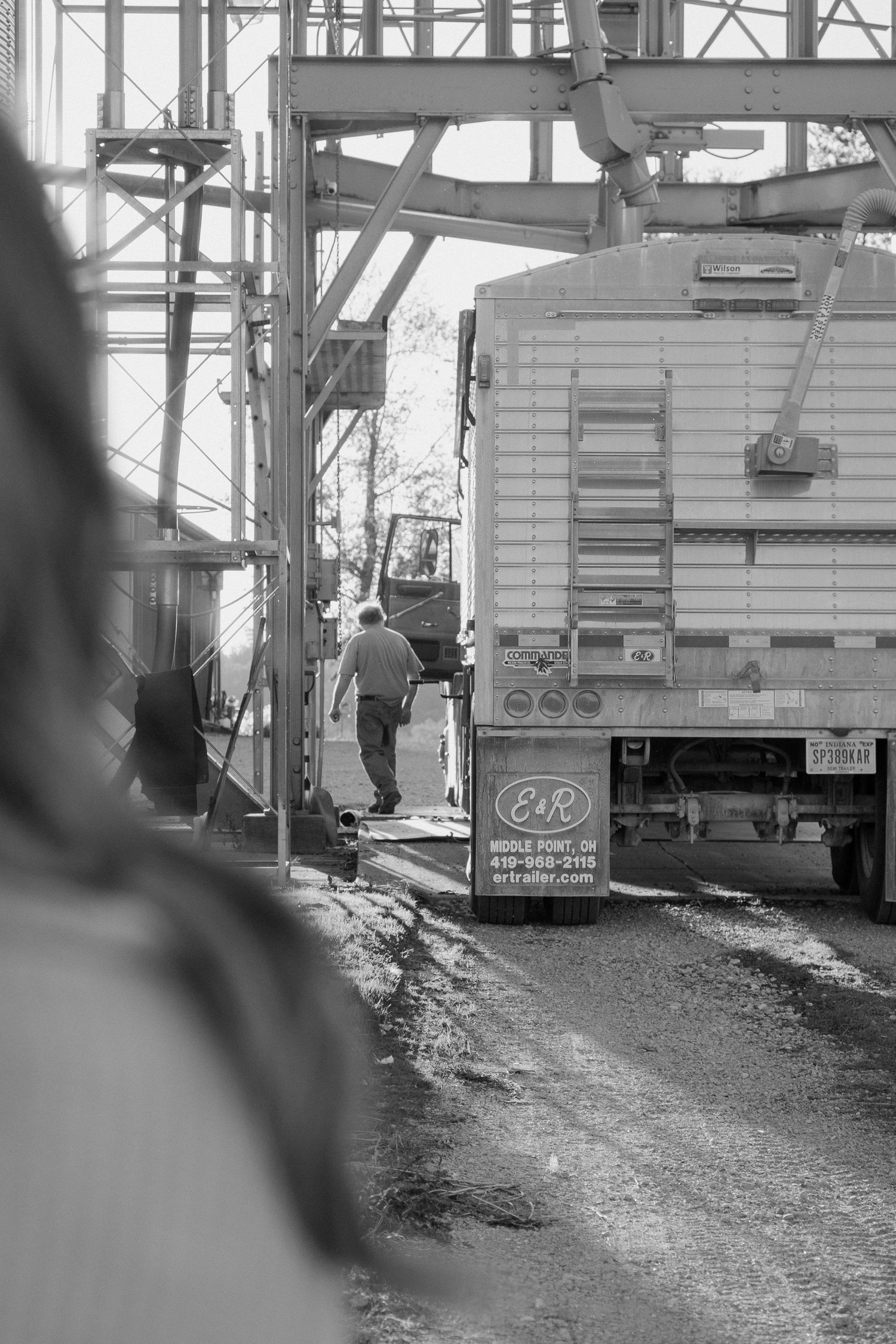 Black and white photo of a farmer walking beside a grain truck at an agricultural facility.