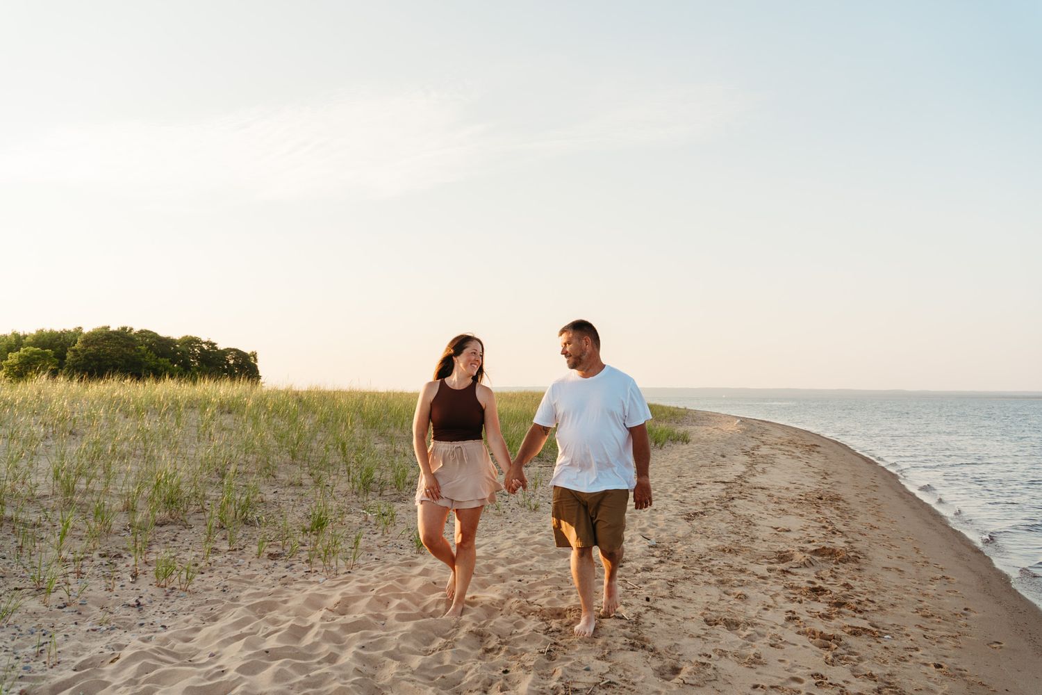 Couple holding hands walking barefoot on sandy beach at sunset with ocean waves and green dunes in background.