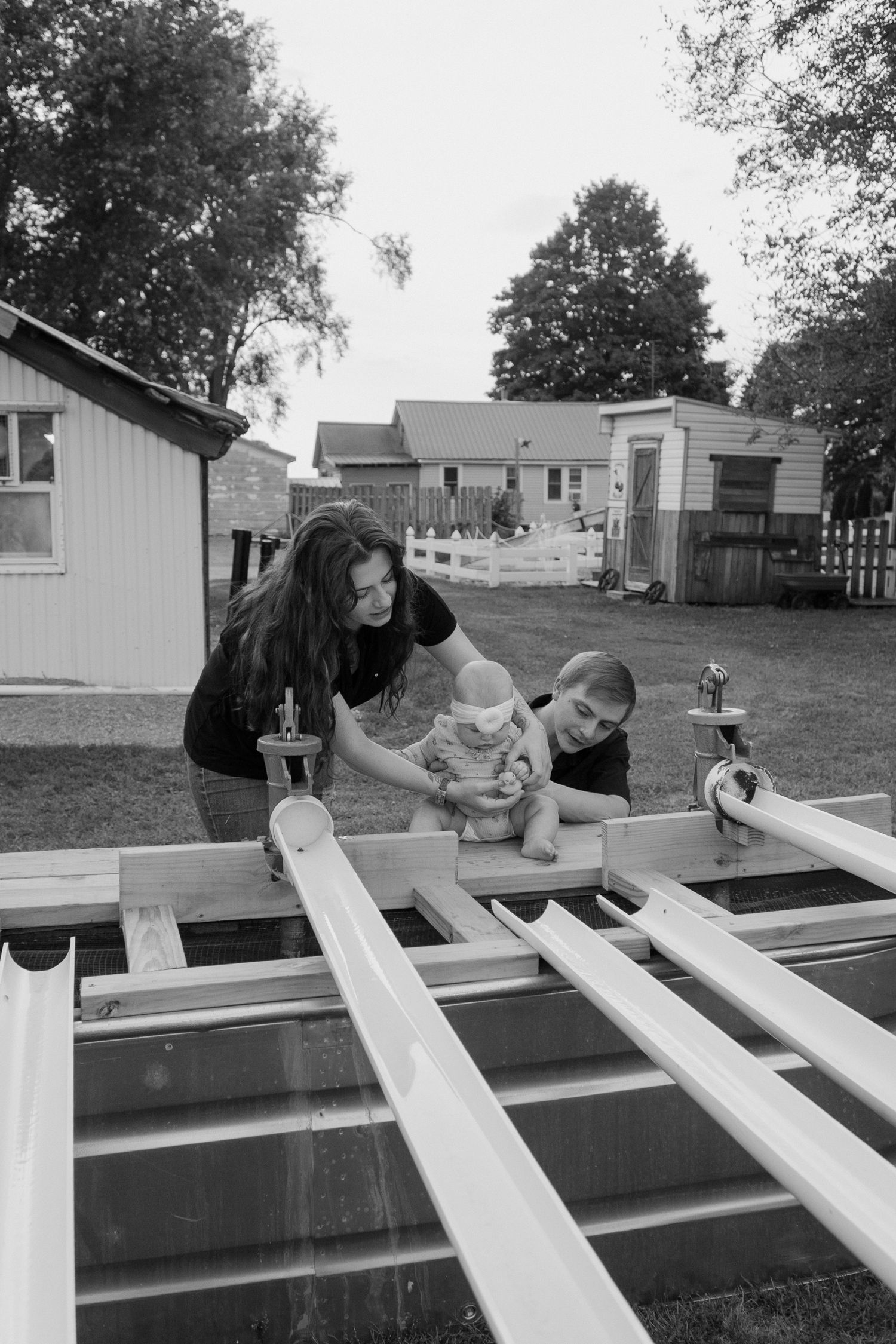 Black and white photo of a couple with a baby playing on a white wooden ladder in a backyard.