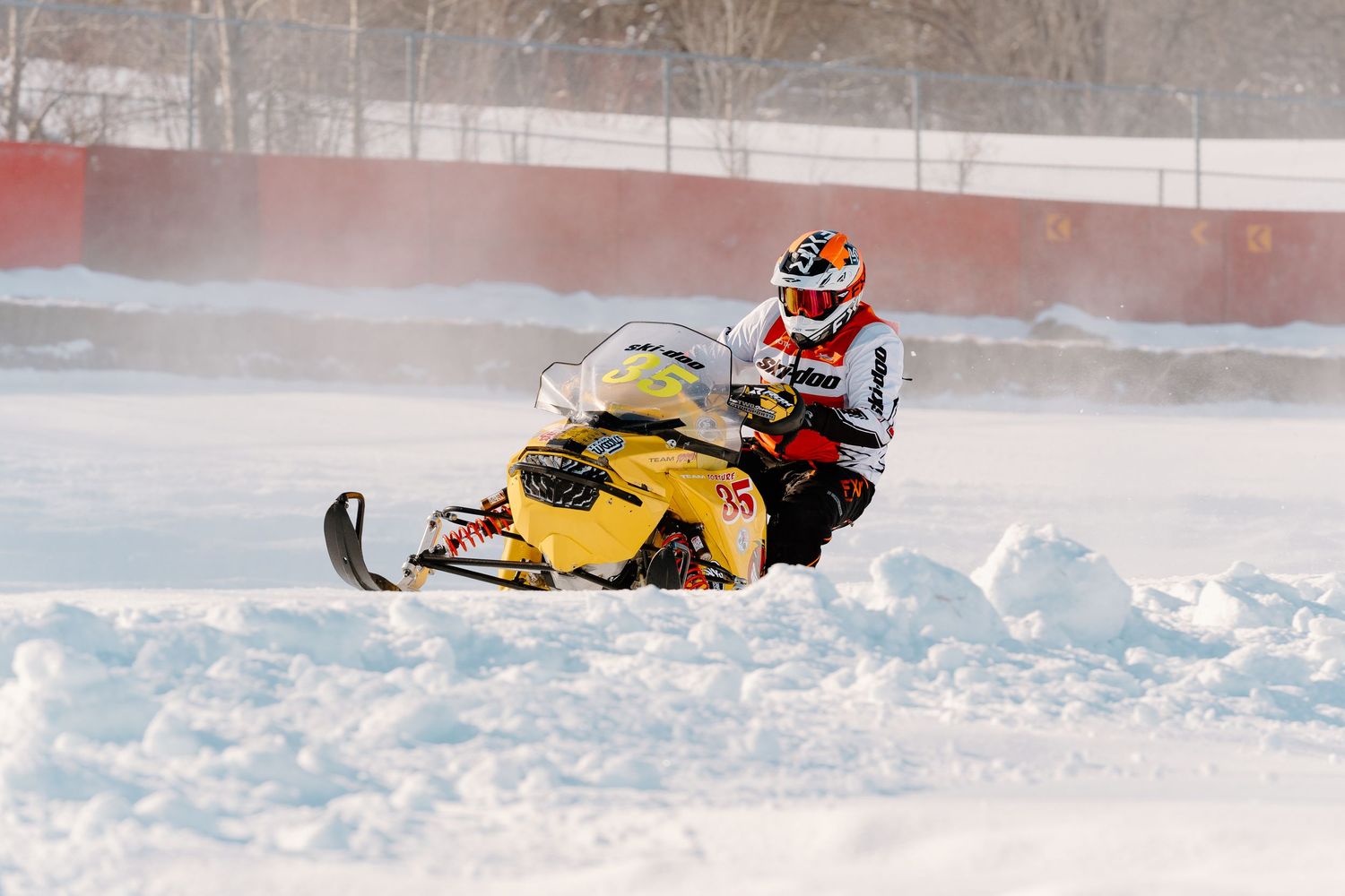 Snowmobile racer in red and white helmet racing a yellow sled through deep snow at high speed.