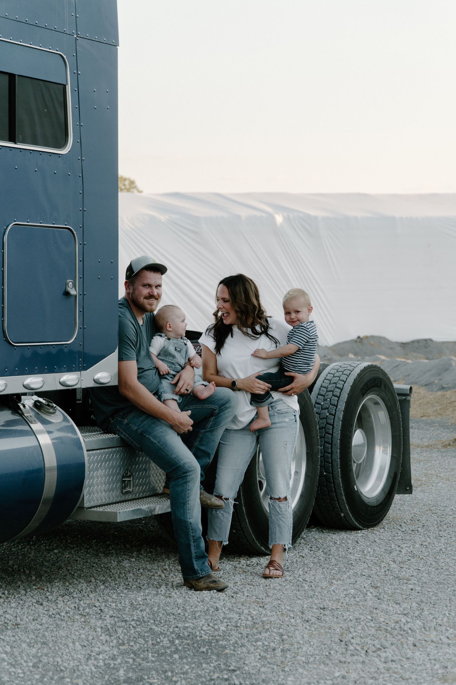Young family with two babies posing beside a blue semi truck on a gravel lot at dusk.