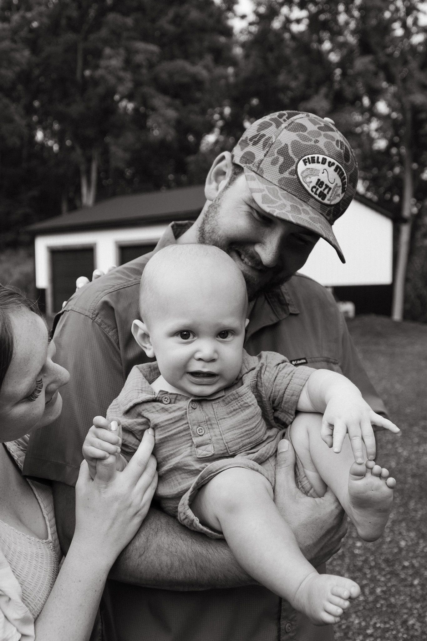 Black and white photo of parents lovingly holding smiling baby outdoors in natural setting.