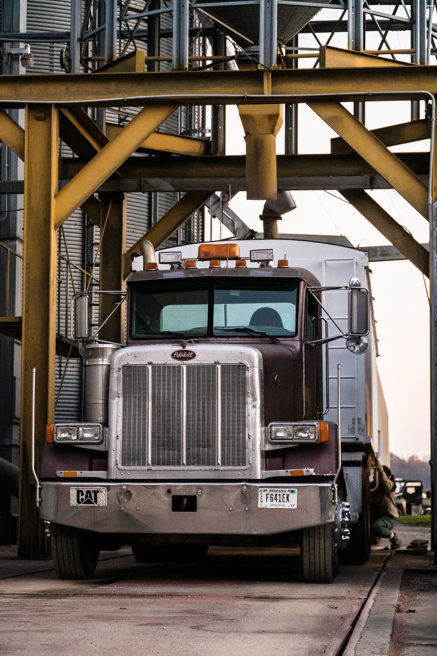 Maroon Peterbilt semi-truck parked under grain elevator structure at agricultural facility during harvest season.