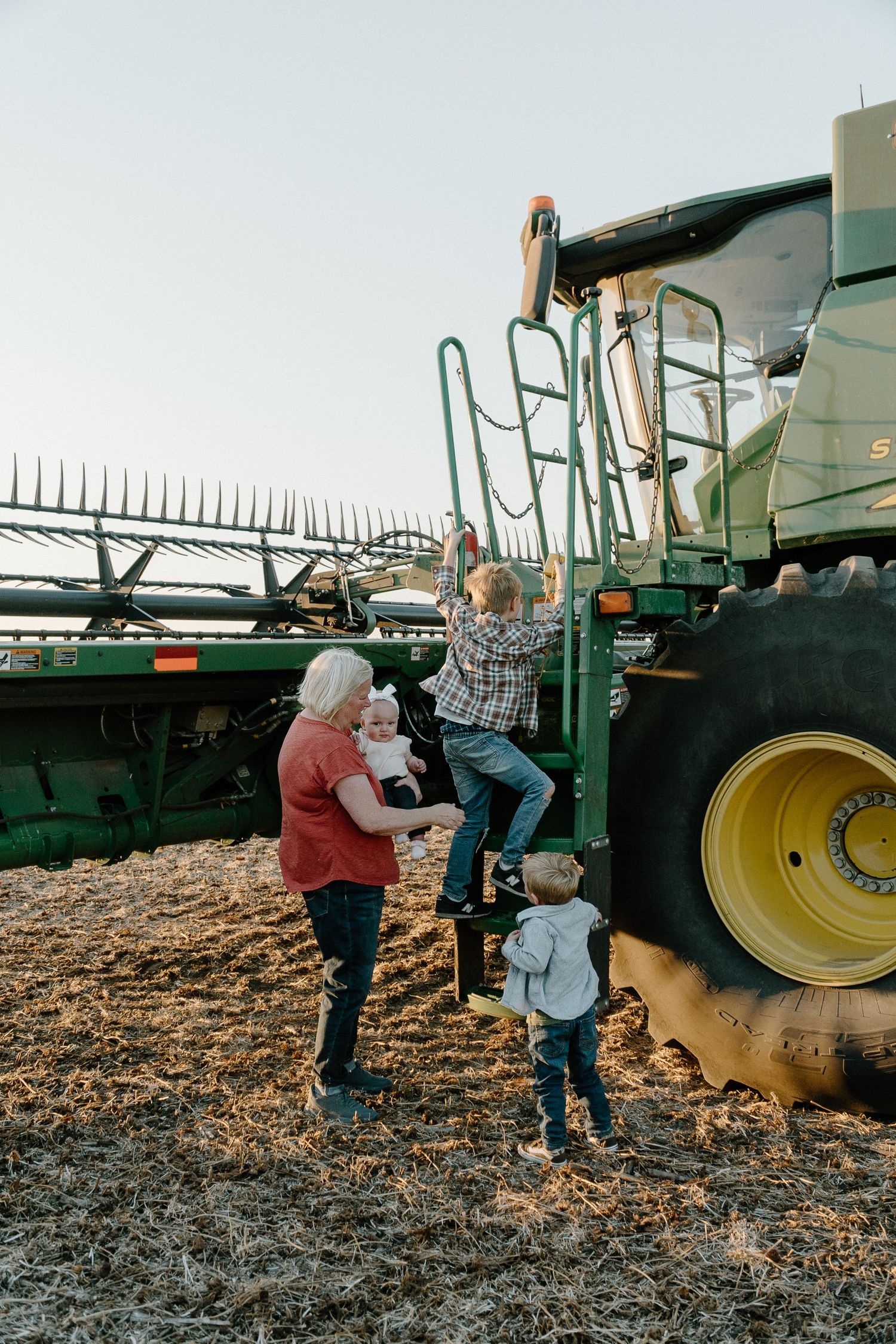 Grandmother with grandchildren climbing a green John Deere combine harvester in a harvested field.