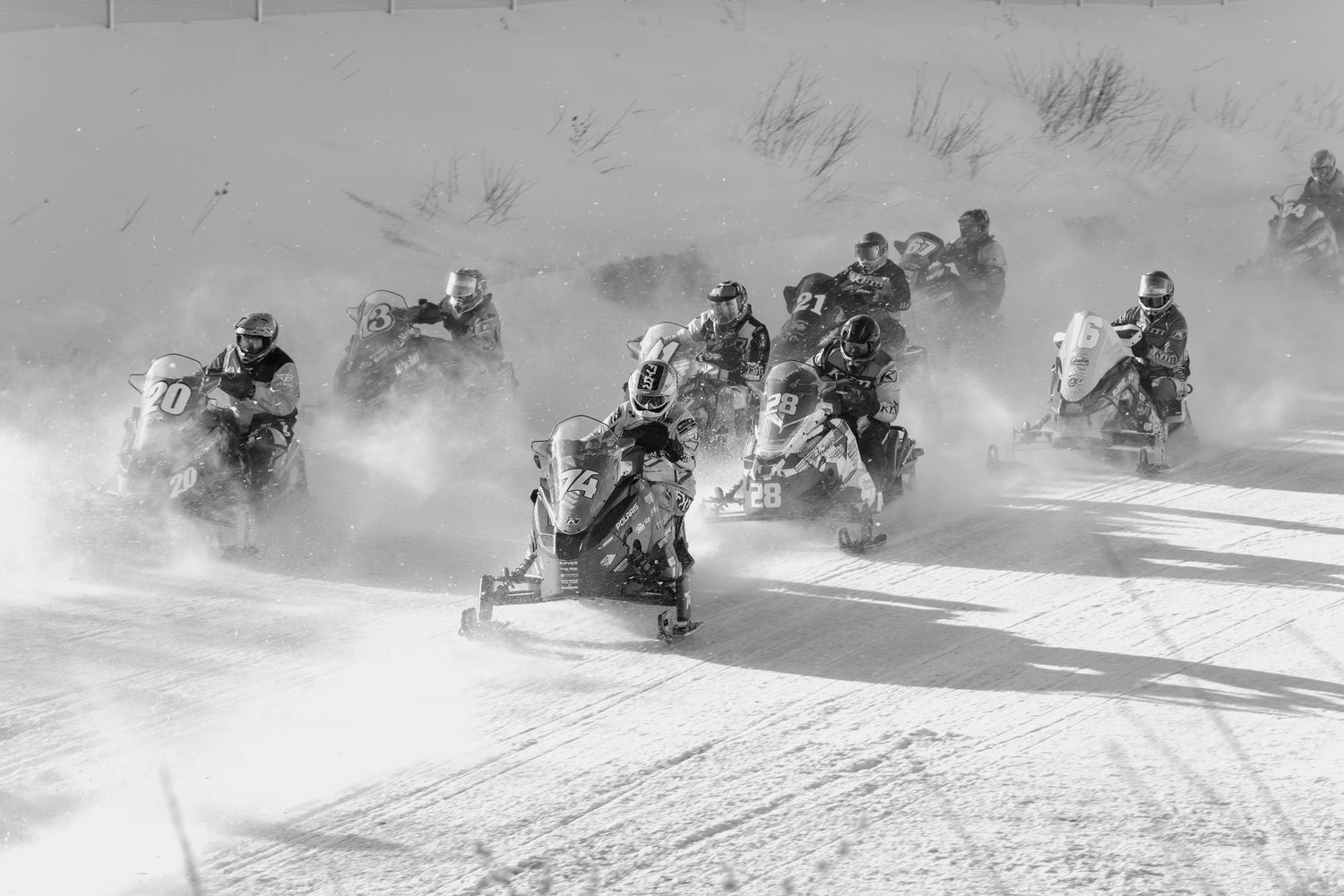 Black and white action photo of multiple snowmobile racers competing on a snowy race track in winter.