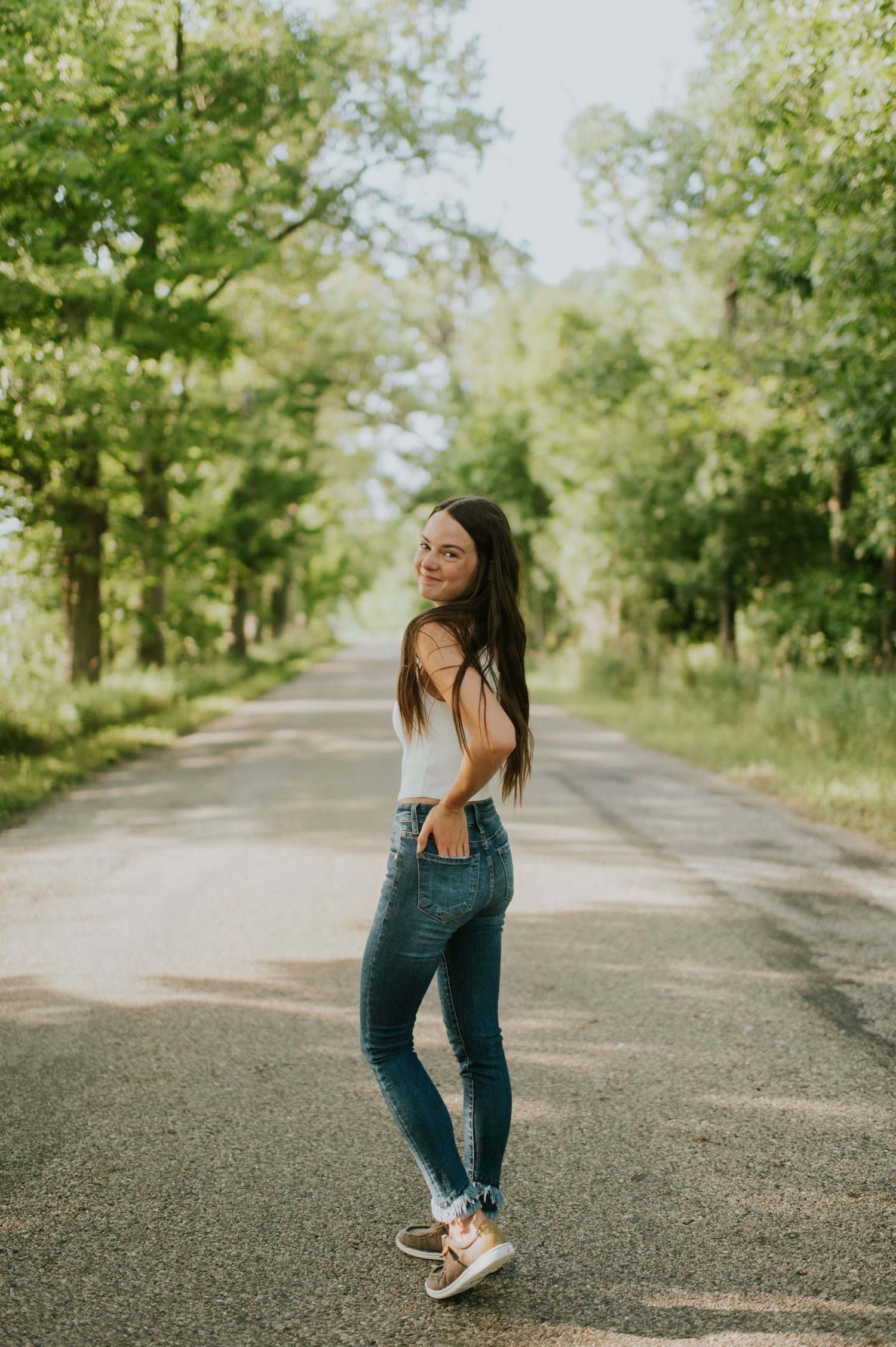 Young woman in white tank top and jeans posing on a tree-lined road in sunlight.